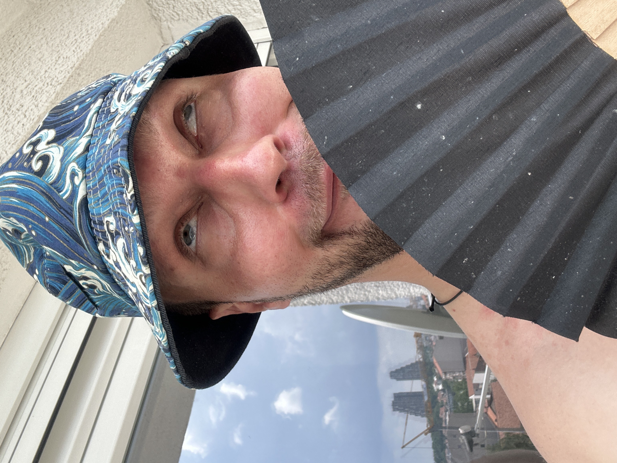 A close-up selfie shows a man wearing a blue and white patterned bucket hat. He is holding a black fan in front of his face, obscuring his mouth and chin. His eyes are looking upwards and to the right. The man has a fair complexion and stubble on his face. He is shirtless, with his shoulder and upper arm visible. In the background, a cityscape is visible under a cloudy sky. The image appears to have been taken on a balcony, with horizontal blinds in the upper left corner and a textured wall to the right. The cityscape includes buildings and a construction crane, suggesting an urban environment. The specific location is the Zvezdara Urban Municipality in Serbia, as indicated by the architecture and skyline resembling the area.