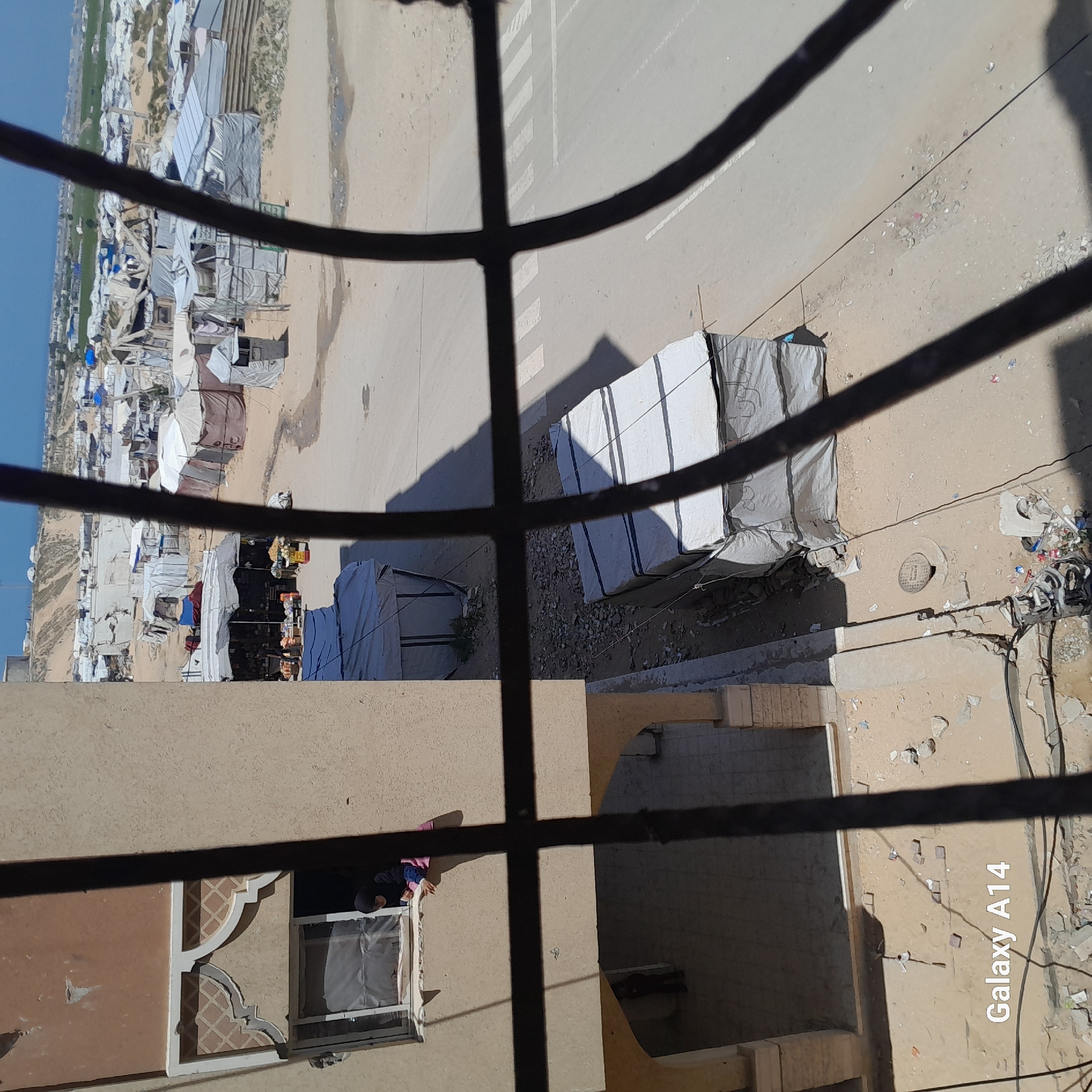 This photograph, taken through a barred window, depicts an urban scene. To the left, a tent city or refugee camp extends into the distance, situated on sandy terrain. In the foreground, a street is visible, with what appears to be a vehicle or structure covered with a white tarp. Debris and trash are scattered along the roadside. The adjacent building has an open window, revealing a person inside. The image is labeled "Galaxy A14." The location is the city of Unknown, Unknown.