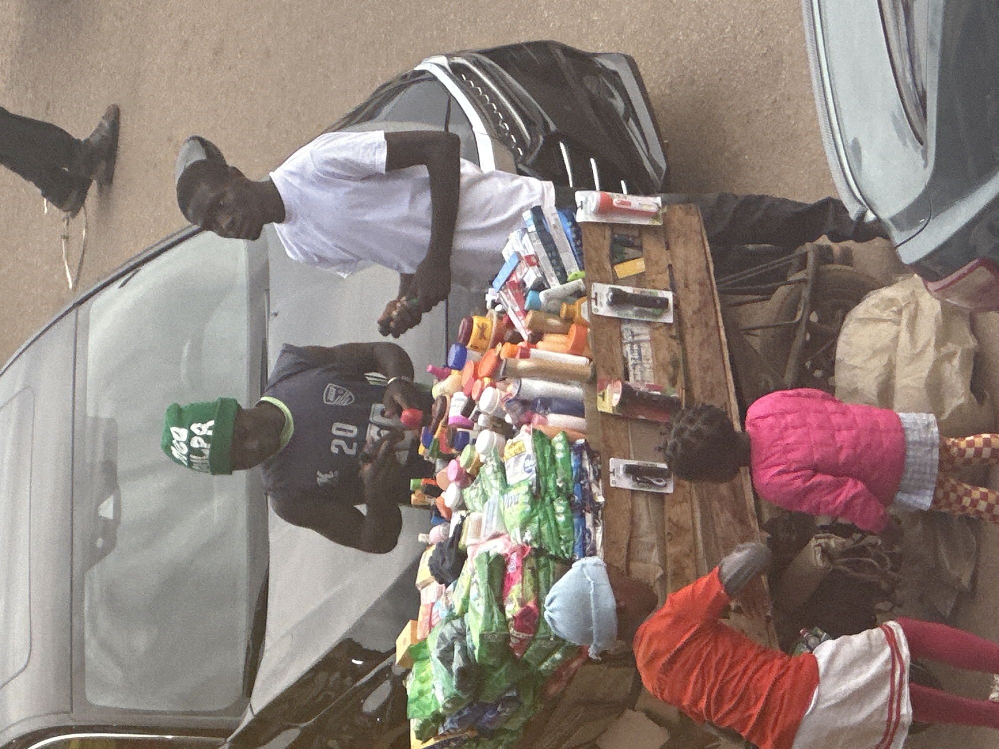 In Bukuru, Nigeria, a street vendor stands behind a stall laden with various goods, including packaged soaps, sanitizers, and other toiletries. The stall is positioned next to a black car, with its trunk open, suggesting the car may be used for transport of goods or as a makeshift display. Two men, likely vendors, are present at the stall. One man, wearing a green beanie and a dark blue t-shirt with the number 20 visible, appears to be interacting with a customer or arranging merchandise. The other man, wearing a white t-shirt, stands slightly behind him, also appearing to be involved in the sale. In the foreground, a young child wearing a pink jacket and checkered pants looks towards the stall, while another child, dressed in an orange shirt and white skirt, bends down near the stall, possibly assisting or browsing. The overall scene depicts a typical street commerce environment in a Nigerian city, with everyday items being sold from a portable stall.