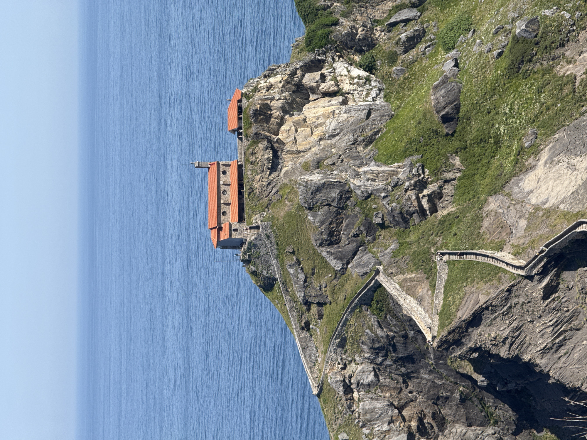 The image depicts a dramatic coastal landscape in Bakio, Spain, featuring a small, ancient-looking church perched atop a rocky island. A winding stone staircase, with numerous steps, ascends the rugged terrain to reach the church, creating a visually striking pathway. The church itself has a red-tiled roof and appears to be constructed of stone, blending harmoniously with its natural surroundings. The deep blue of the ocean stretches out to the horizon, providing a vast and serene backdrop to the scene. The island is covered in patches of green grass and interspersed with rocky outcrops. The sky above is a clear, pale blue, indicating a bright, sunny day. There are a few people visible on the staircase and near the church, suggesting it is a popular tourist or pilgrimage site. The overall impression is one of historical significance and natural beauty, a testament to human resilience and faith against the elements.