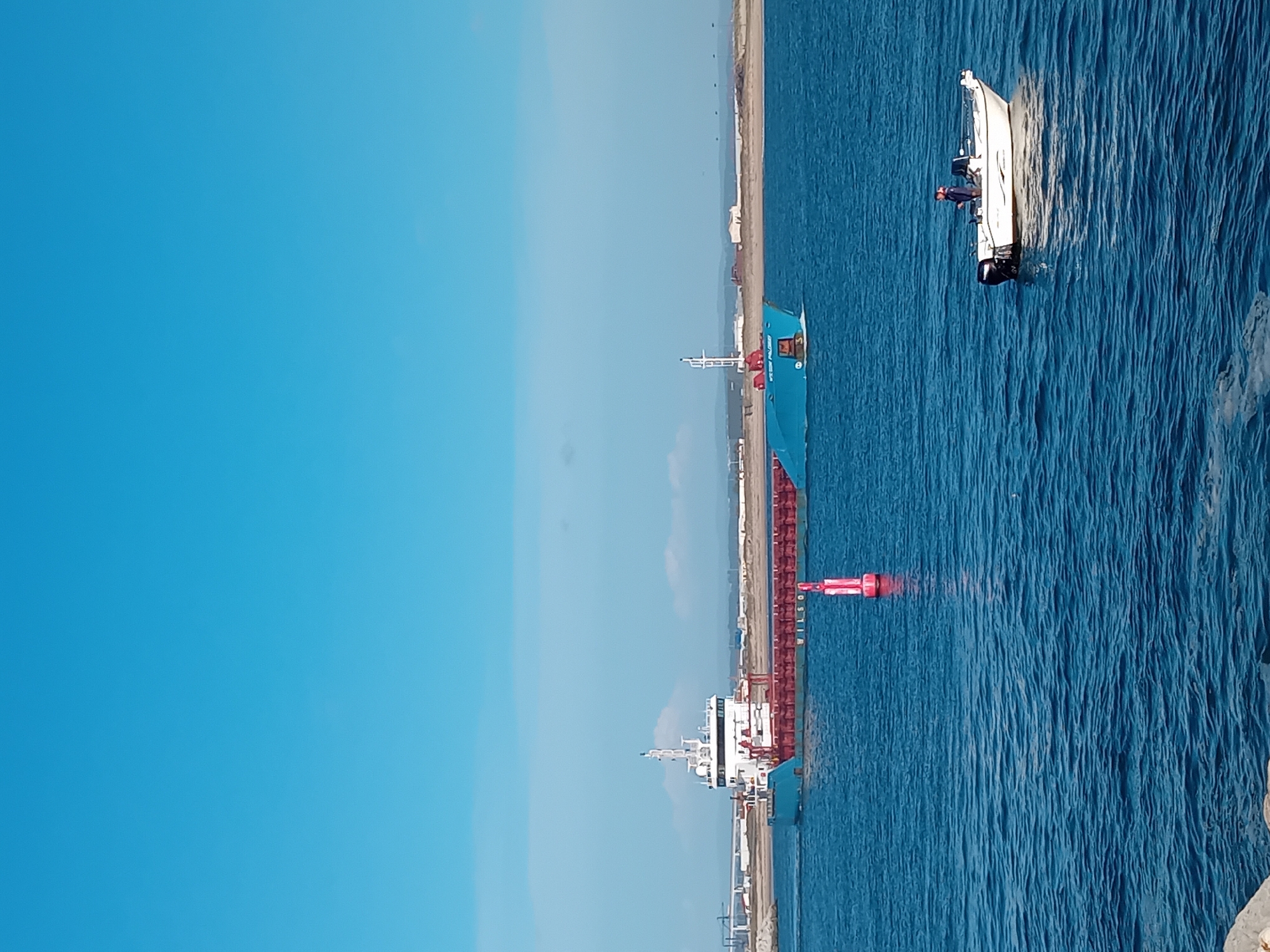 The image shows a cargo ship docked at the Port of Figueira da Foz in Portugal. A smaller boat is near the entrance to the port. The sky is clear and blue. The sea is calm. A person is on the small boat.