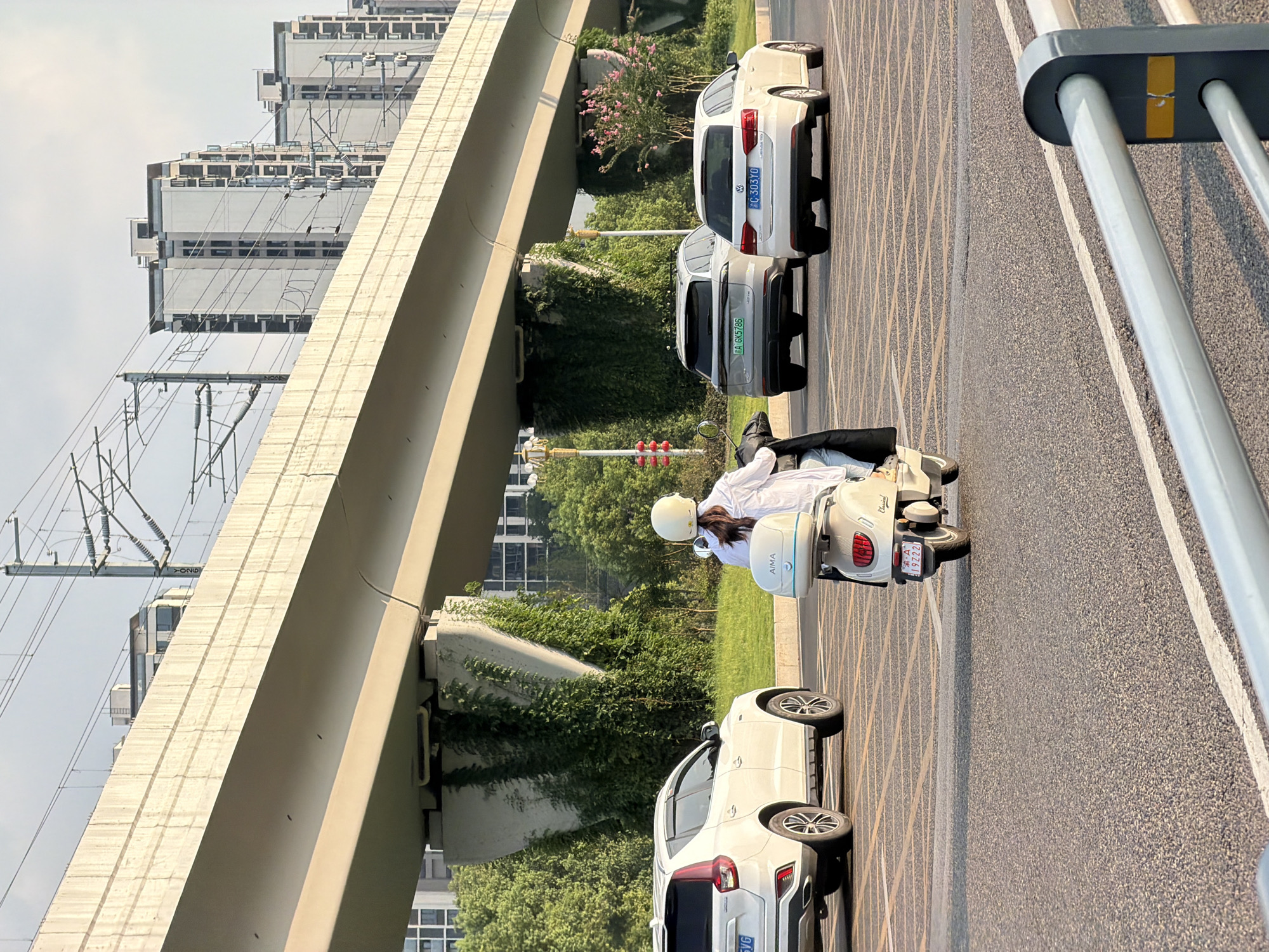 In Jiangjin, China, on a sunny day, a person rides a white electric scooter with a black bag attached to the back, traveling on a road. The rider, wearing a white shirt and white helmet, is positioned in the foreground of the image, facing away from the camera. Ahead of the scooter, two white cars are visible, a sedan in the right lane and an SUV in the left lane. The road surface is asphalt, marked with yellow lines. In the background, an elevated concrete structure, likely a highway or overpass, spans across the scene, supported by pillars. Green foliage, including trees and vines, grows along the pillars and on the sides of the road. Beyond the elevated structure, several tall residential buildings are visible against a clear sky, with overhead power lines and railway tracks present. The scene captures a moment of urban transportation with a blend of modern infrastructure and natural elements.