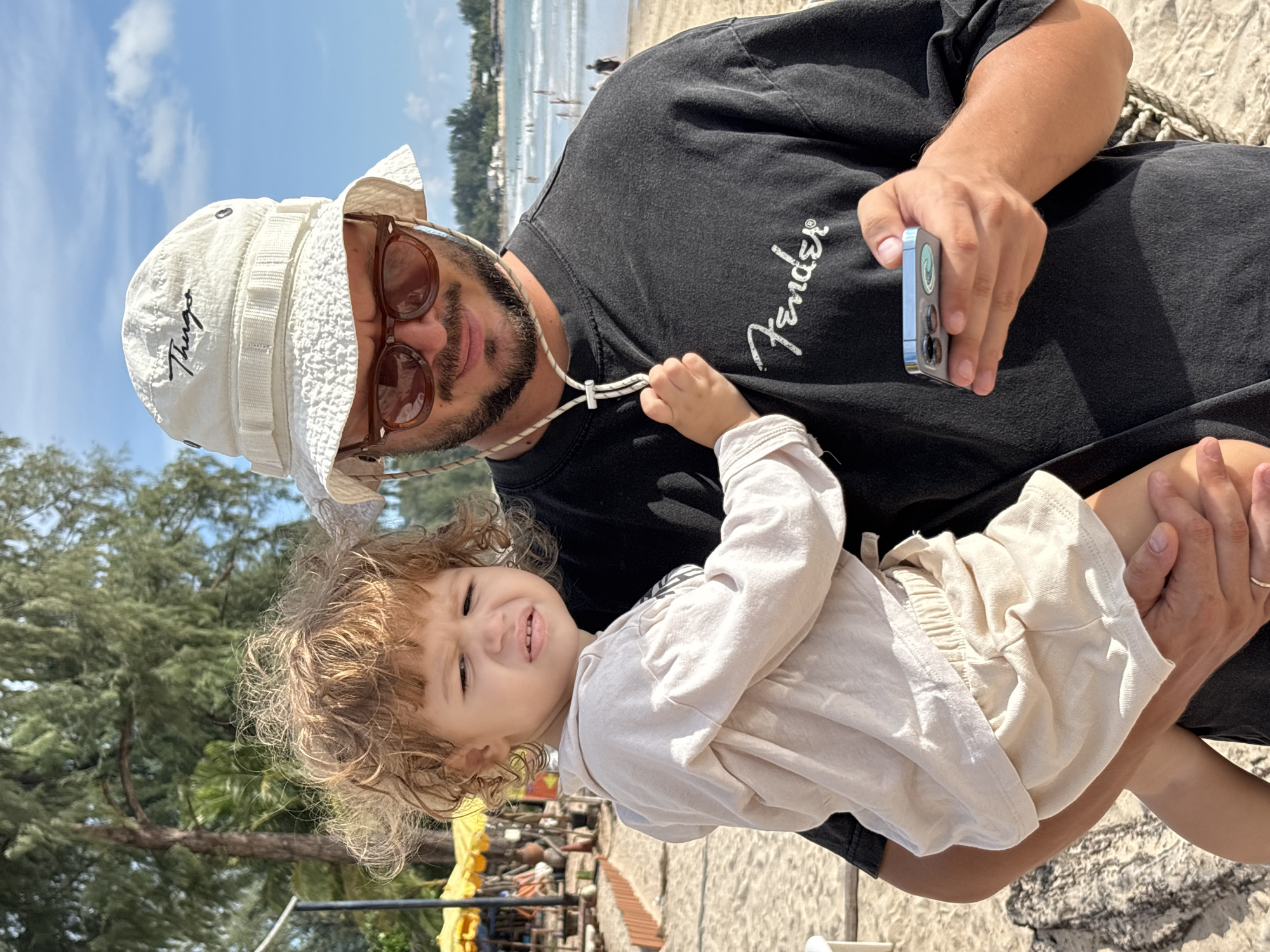 A man wearing a white bucket hat and sunglasses holds a young child on a beach in Choeng Thale, Thailand. The child, with curly blonde hair, wears a light-colored shirt and shorts and has a distressed expression. The man, dressed in a black t-shirt with a "Fender" logo, is looking at his phone, which he holds in his right hand. In the background, trees and beach umbrellas are visible, along with people in the ocean and on the sand. The sky is blue with scattered clouds.