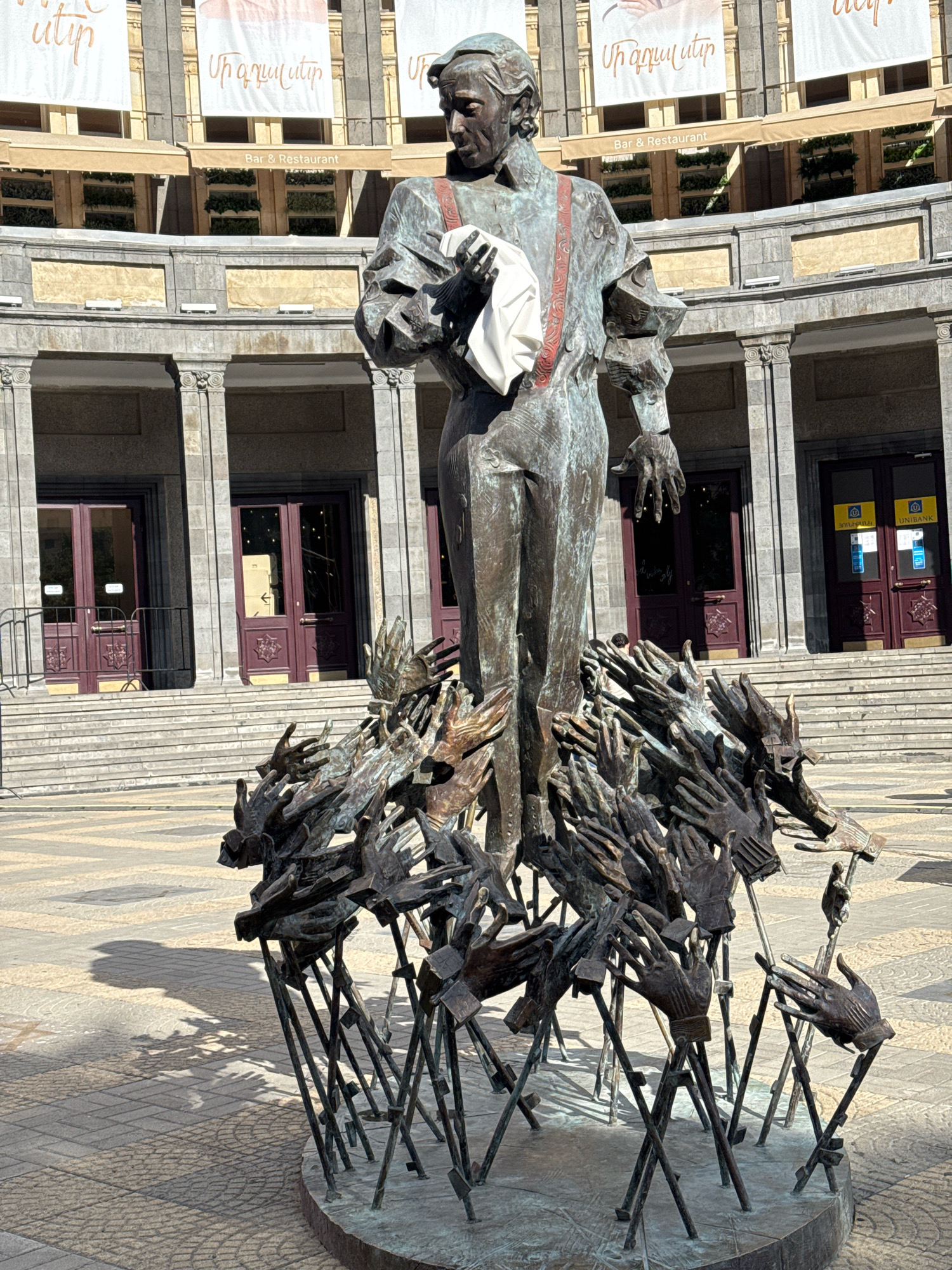 The image shows a bronze statue of a man standing on a base of many outstretched hands in front of a building. The statue depicts a man in historical clothing, holding a white cloth in his left hand. He wears red suspenders over a white shirt and dark pants. The hands at the base of the statue are also made of bronze and are arranged in a chaotic, upward-reaching pattern, attached to thin metal rods. The building in the background has classical architectural elements, including columns and a grand entrance with stairs. Banners with text hang across the facade of the building. The ground in front of the statue is paved with square stones, and sunlight casts shadows. This statue is located in front of the Opera and Ballet Theatre in Yerevan, Armenia.