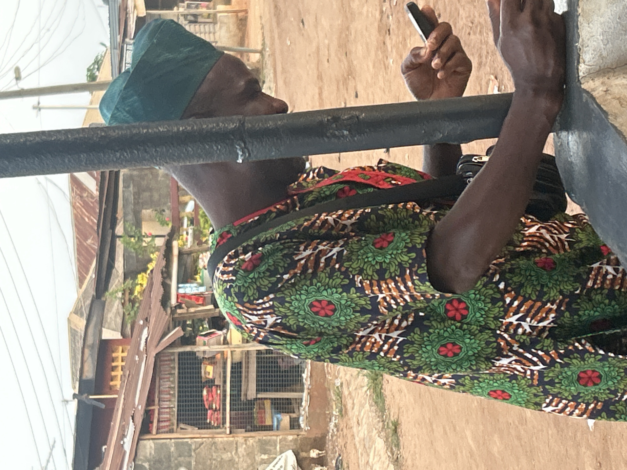 The image shows a person wearing a patterned green and red shirt and a green head covering, holding a mobile phone in their left hand. They are leaning on a black metal bar or railing. A black strap of what appears to be a bag or camera is slung over their shoulder. The background shows buildings, one with a corrugated metal roof, and an apparent storefront. The scene is set in an urban environment, with a visible street or pathway.