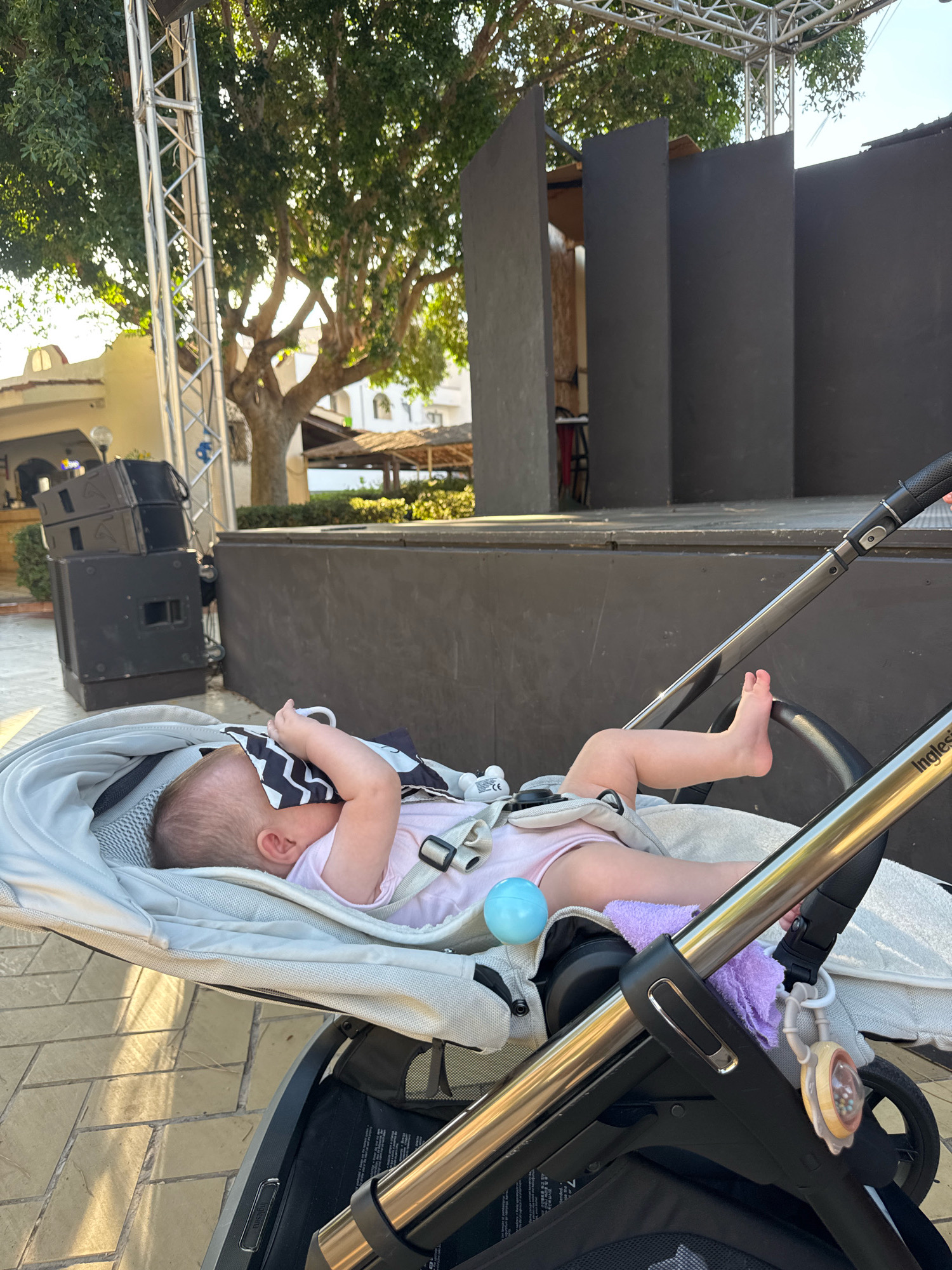 An infant lies asleep in a stroller outdoors during daylight. The baby is wearing a pink onesie and is partially covered by a blanket with a black and white zig-zag pattern, which they are holding over their face. The stroller is a neutral color and has a metallic frame. A small blue ball is visible near the baby's torso. In the background, there is a stage with dark panels and scaffolding, suggesting an outdoor event or performance space. Trees with lush green foliage are also present, and sunlight filters through them. The ground is paved with rectangular stones. The scene appears to be in Isola delle Femmine, Italy, possibly near a venue or recreational area.