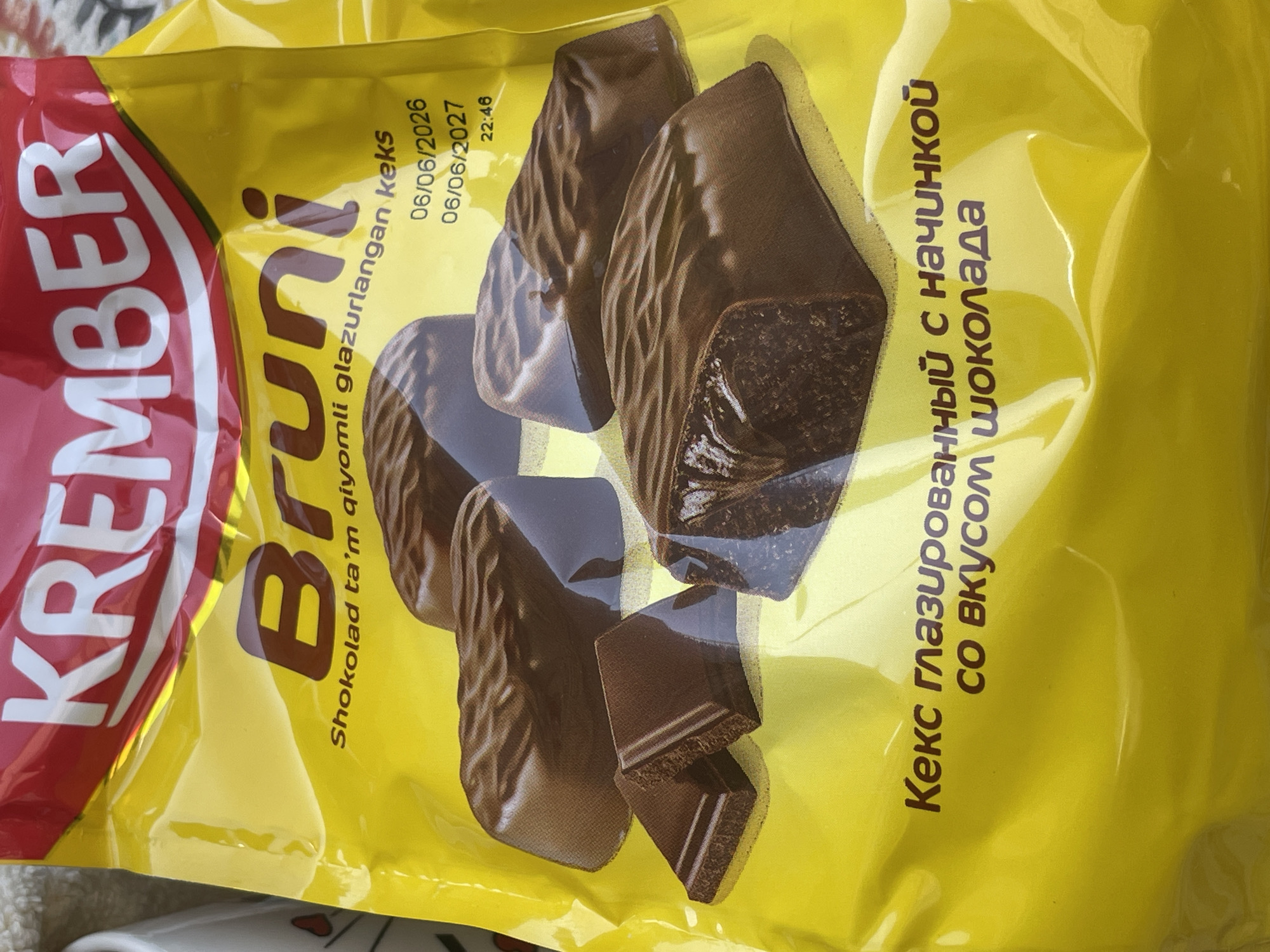 The image shows the packaging of "Bruni" brand chocolate-covered cakes. The packaging is primarily yellow with a red logo at the top that reads "KREMBER". Below the logo, "Bruni" is written in large brown letters, followed by smaller text in Uzbek that translates to "chocolate-flavored glazed cakes". There are images of chocolate cakes on the packaging. In the bottom portion of the packaging, there is text in Russian that reads "Кекс глазированный с начинкой со вкусом шоколада", which translates to "Glazed cake with chocolate filling". The expiration dates on the package are "06/06/2026" and "06/06/2027". The image does not provide any information about a specific location in Tashkent, Uzbekistan, or any details about people, actions, or interactions.
