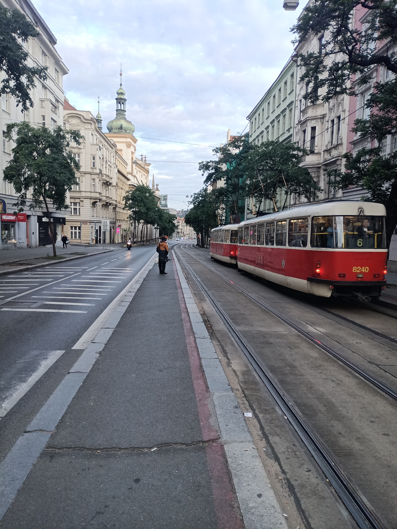 A person wearing a dark jacket and a yellow-orange backpack stands on a sidewalk next to tram tracks. The person is looking away from the camera. A red and white tram with the number 8240 on its front is on the tracks to the right. The tram has several windows, some of which are occupied by passengers. The tram is long, appearing to be two connected sections. The street is paved with asphalt, and there are white markings on the road indicating a crosswalk. Buildings line the street on both sides, some with ornate architectural details and domed roofs. Trees with green foliage are planted along the sidewalk and in front of the buildings. The sky is overcast and the lighting suggests it is either early morning or late afternoon. The scene depicts an urban street with active public transportation and pedestrian activity in Prague, Czechia.