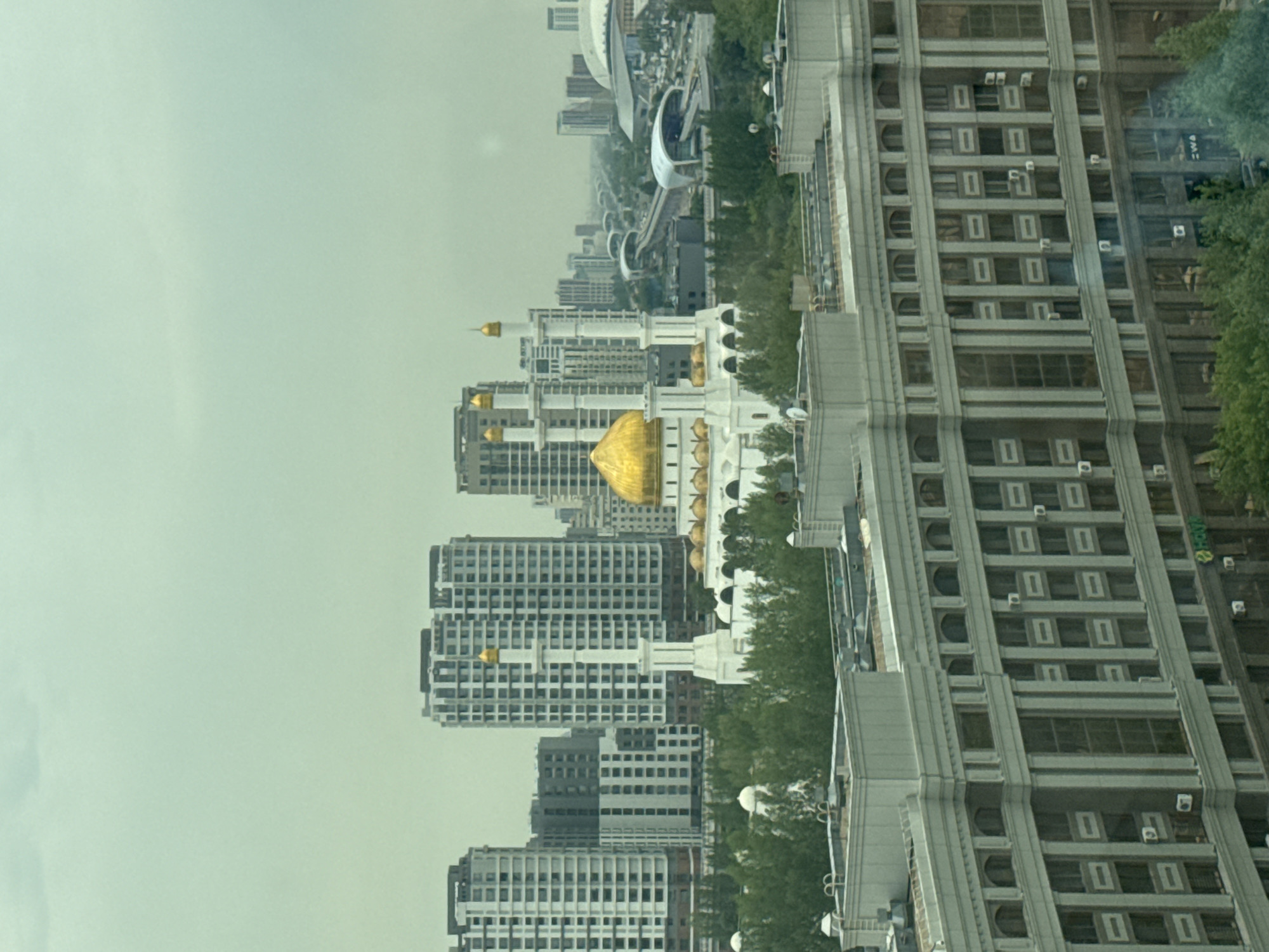 The image depicts a panoramic view of Astana, Kazakhstan, under an overcast sky. In the foreground, a large, modern, light-colored building with multiple floors and numerous windows is visible. This building has a distinct architectural style, featuring columns and a detailed facade. Behind it, a lush green canopy of trees forms a natural barrier.
Dominating the mid-ground is the iconic Hazrat Sultan Mosque, a grand white structure with a large golden dome at its center and several smaller golden domes. Two tall, slender minarets with golden tips stand prominently beside the mosque. The mosque is surrounded by trees and is part of a cityscape that includes several contemporary high-rise buildings with varied designs and many windows.
Further in the background, the urban landscape of Astana unfolds, showing a network of modern buildings, elevated train tracks with futuristic-looking stations, and various other structures stretching into the distance. The overall impression is of a modern, developing city with a blend of architectural styles, nestled amidst a green environment, under a muted, cloudy sky.