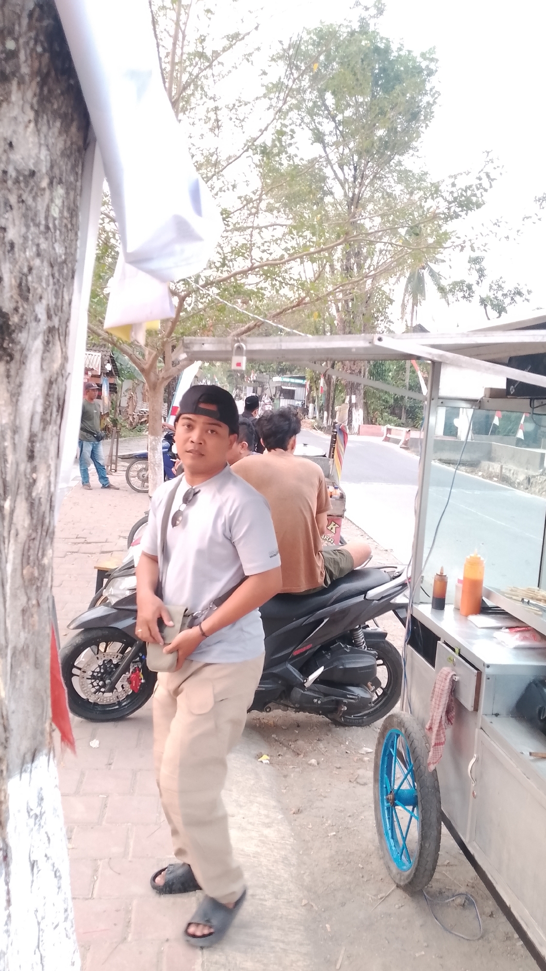 The image depicts a street scene in Karanglangu, Indonesia. In the foreground, a man wearing a black cap, a grey t-shirt, and beige cargo pants stands next to a black motorcycle. He has a small beige bag slung across his chest and sunglasses hanging from his t-shirt collar. To his right, a food cart with a blue wheel is visible, containing bottles of sauces and skewers of food. In the background, another person is seated on the motorcycle, wearing a brown t-shirt and green shorts. Further down the street, other individuals and various structures, including trees and a building with awnings, are present. The scene appears to be a casual outdoor setting, likely a roadside food stall.
