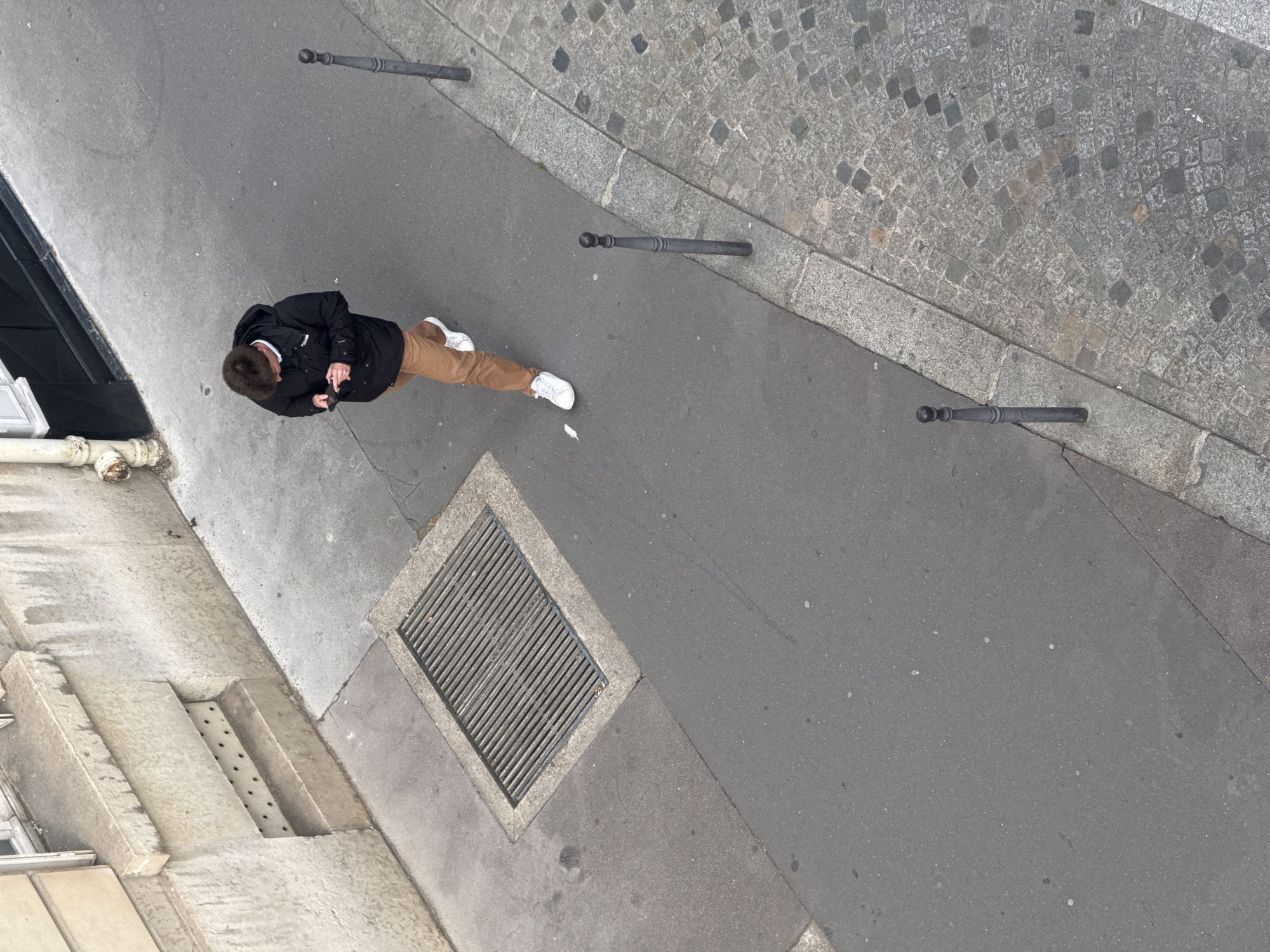 The image is a top-down shot taken in Paris, France. A person wearing a black jacket, brown pants, and white sneakers is walking on a gray asphalt sidewalk. The person appears to be looking at a phone in their hands. The sidewalk is bordered by a cobblestone street and features short, cylindrical metal bollards. A metal grate is embedded in the sidewalk. A building facade is visible on the left side of the frame.