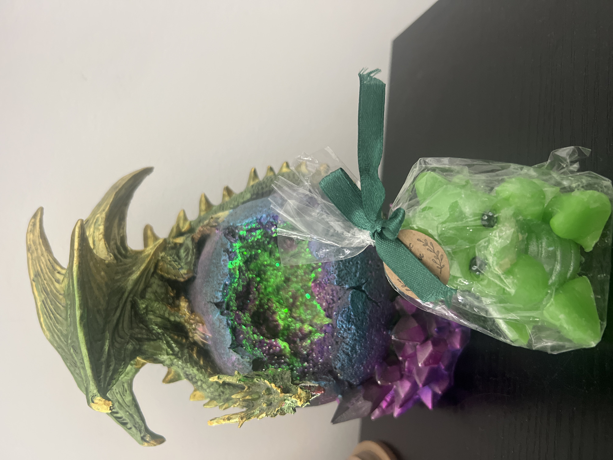 On a dark surface next to a white wall is a dragon figurine and a plastic-wrapped gift. The dragon is painted green, gold, purple, and blue. The dragon's head and wings are on the left. The body is covered in iridescent paint and crystals. Next to the dragon is a clear plastic bag tied with a green ribbon and a brown tag. Inside the bag are green candy or soap shaped like hearts. The location is not specified in the image. Therefore, the request for the location is impossible.