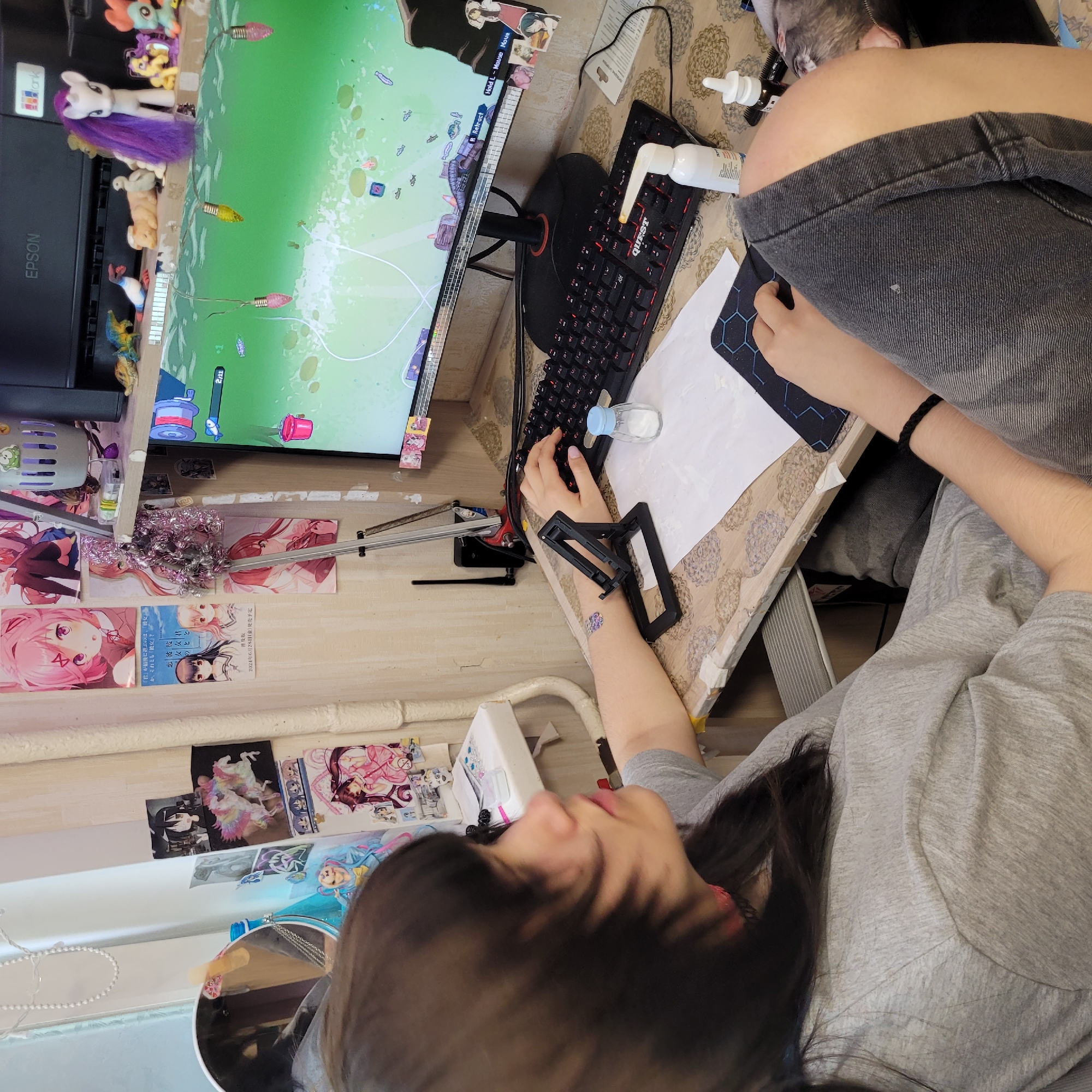 A high-angle shot captures a person absorbed in playing a video game at a desk within what appears to be a home setting. The person, with long dark hair, wears a grey t-shirt and dark shorts, her face partially obscured as she looks down at the monitor. Her right hand is positioned over a black "QUEST" brand keyboard, while her left hand rests on a dark hexagonal mousepad, operating the mouse. The computer monitor displays a brightly colored game, possibly a fishing simulator, featuring a green aquatic environment with fish, a fishing reel, and a line. Visible game text includes "Hold L-Mouse Move" and "R Retract," along with a "2/11" counter.
The desk surface is covered with a patterned mat and holds various items, including a black phone stand, a white piece of paper, a white nasal spray bottle, and a small salt shaker with a blue lid. To the left of the monitor, an "EPSON" printer is visible, topped with several My Little Pony figurines, notably a white pony with purple hair.
The wall space behind the desk is decorated with numerous anime-style posters and pictures. One prominent poster features Japanese text that translates to "Who will 'you' ultimately choose, 'her' or 'her'?" and indicates a release date of "Friday, June 28, 2024." Other text includes "普及版" (Standard Edition) and "恋彼" (Love Boyfriend/Girlfriend). A small circular mirror and other decorative objects also adorn the space. A furry, grey animal, likely a cat, is partially visible in the upper right background, resting near the person's shoulder or lap. The scene conveys a personalized and engaging gaming environment.