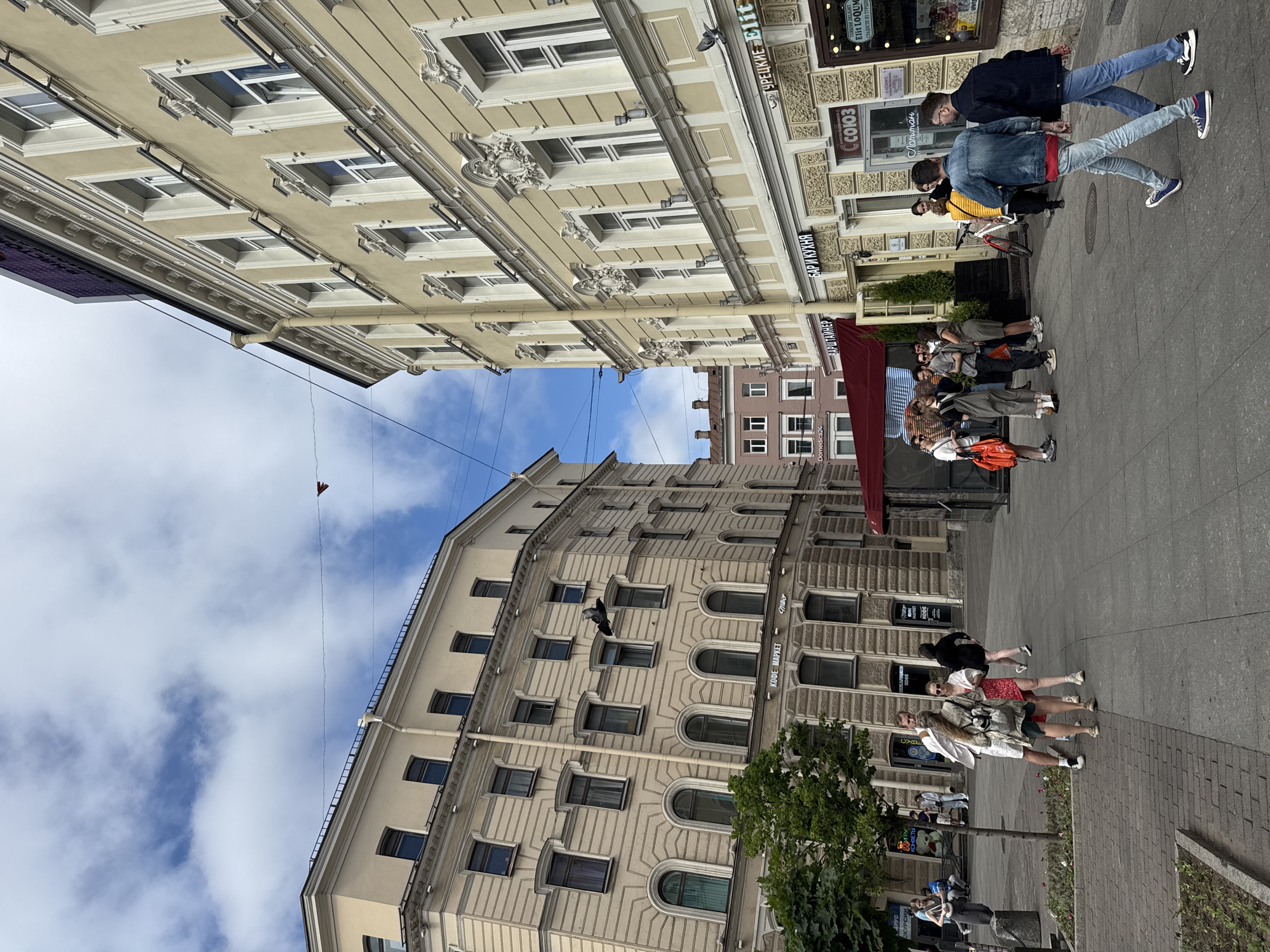 The image shows a street scene in a city, with pedestrians walking on a paved sidewalk. Two large buildings, one on the left and one on the right, line the street. The building on the left is light brown with multiple windows and architectural details. The building on the right is a pale yellow color, also with many windows and ornate decorations. There are some trees on the left side of the image. Several people are scattered across the sidewalk, some walking in pairs or small groups, others alone. One group of people is standing near the center, looking at something or conversing. There is a red awning extending from a building towards the street. Signs with Russian text are visible on the buildings. The sky is partly cloudy.