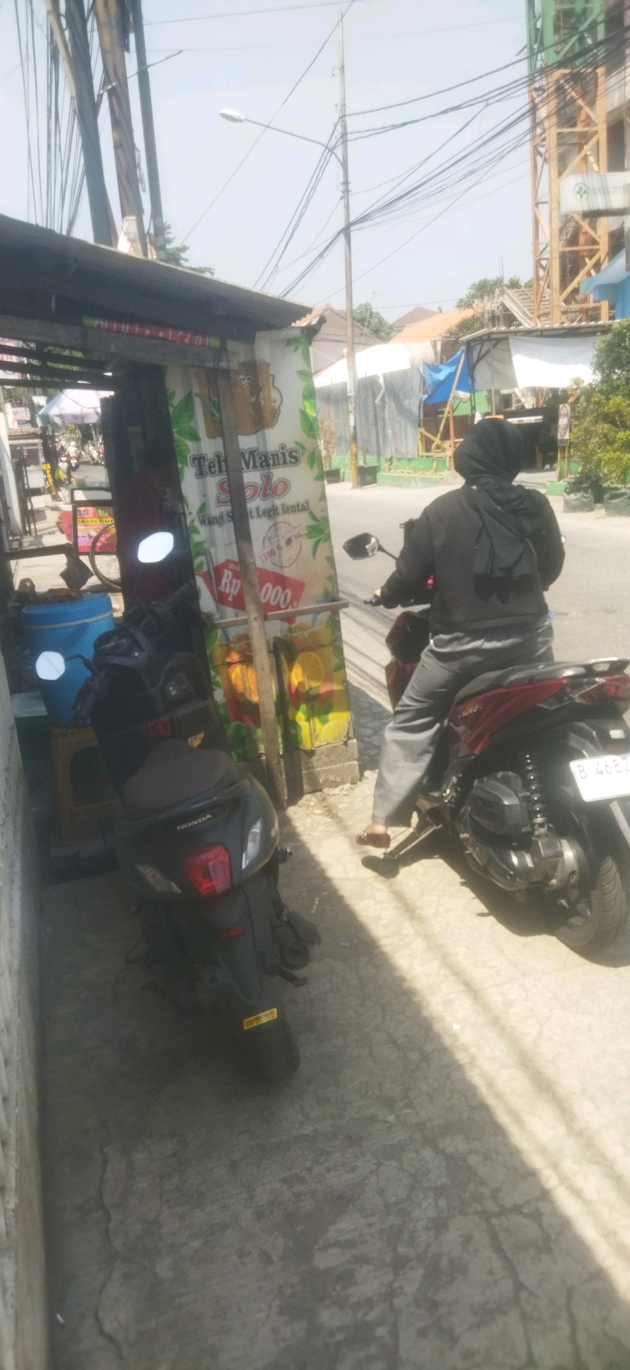 On a sunny day in Depok, Indonesia, a person wearing a black hijab and gray pants rides a red motorcycle away from the camera. The motorcycle's license plate reads "B 4682 Z". The motorcycle is parked on a sidewalk next to a small roadside stall. The stall has a large sign that advertises "Teh Manis Solo" (Sweet Tea Solo) and shows prices in Indonesian Rupiah. Next to the stall, a black Honda scooter is parked. The environment includes a clear sky, overhead power lines, and buildings in the background, one of which appears to be under construction. The ground is paved with concrete.