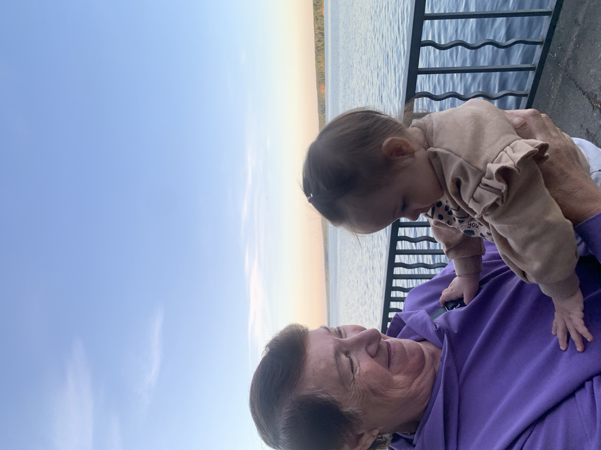 In Chaikovsky, Russia, an elderly woman in a purple hoodie holds a baby in a tan outfit with ruffled sleeves. They are by a body of water, likely a river or lake, with a forested shoreline in the distance. A black metal railing separates them from the water. The sky is a clear blue with a few wispy clouds, suggesting it is either sunrise or sunset. The woman is smiling and looking down at the baby, who is looking away from the camera. The baby's hand is touching the woman's chest.