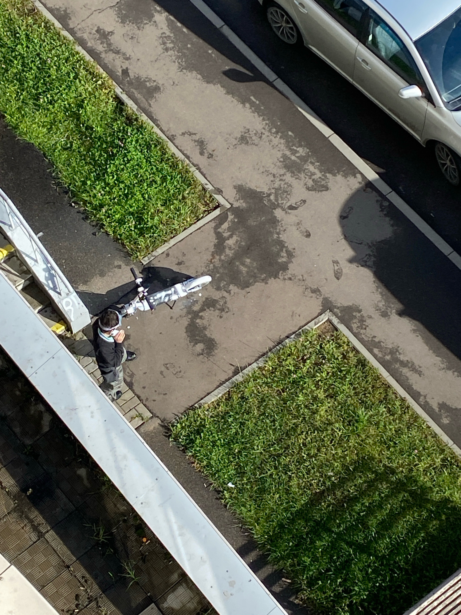 An overhead drone shot captures a street scene from an elevated perspective. A silver station wagon is parked at an angle on a paved surface. To the left of the vehicle, a patch of green grass is visible, bordered by a concrete edge. In the foreground, a person wearing a dark jacket and camouflage pants stands next to a white electric scooter. The scooter has a black and white striped handlebar. The person is wearing a white band around their head and holding something to their mouth. A white metallic structure with yellow accents runs diagonally across the lower left of the frame, suggesting a building facade or balcony. The ground is paved with asphalt and some areas appear wet, with faint footprints visible in the wet patches. Sunlight casts shadows across the scene, indicating it is daytime.