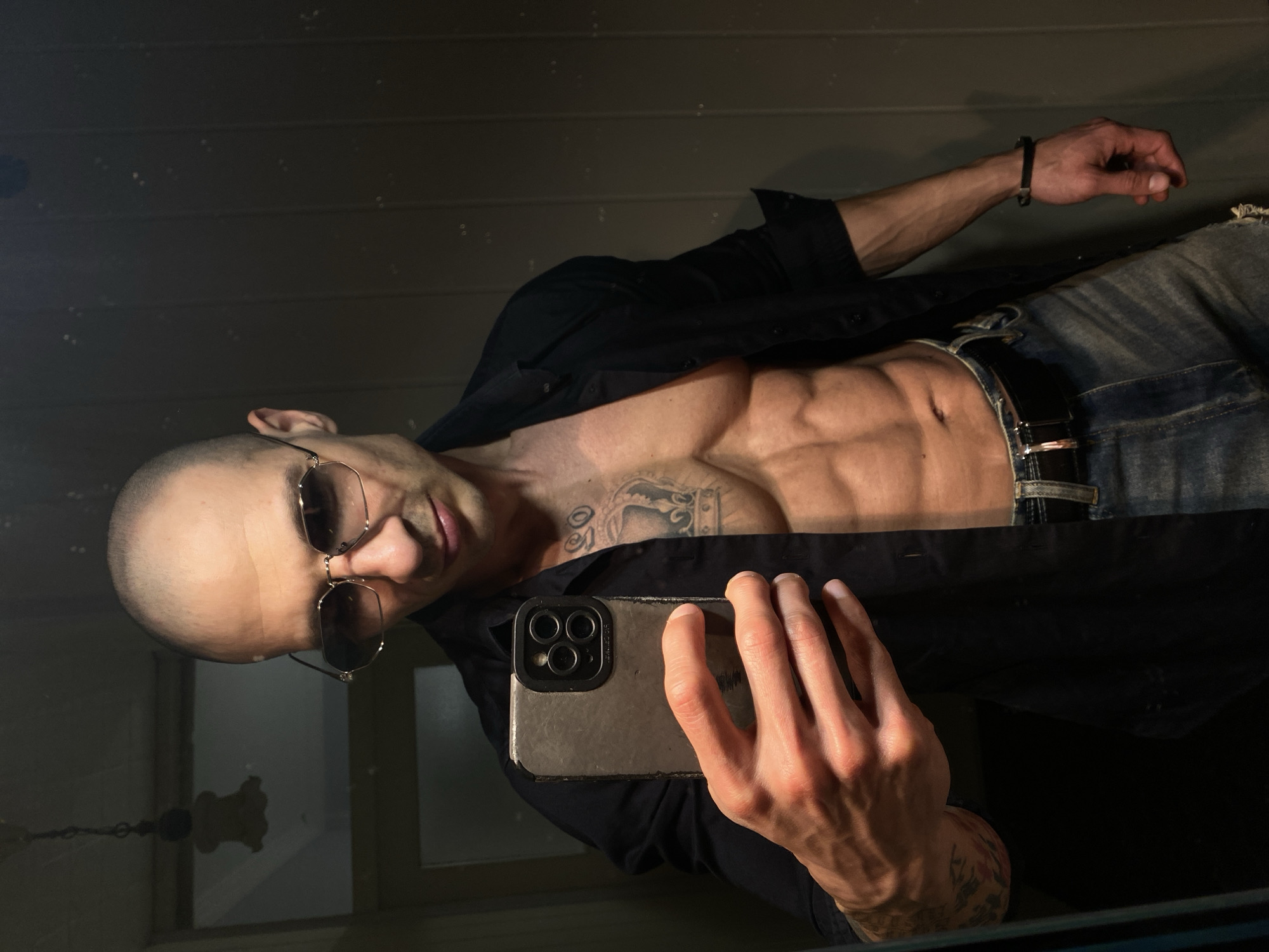 An indoor mirror selfie depicts a bald, muscular male subject, approximately 30-40 years old, wearing mirrored hexagonal sunglasses and an unbuttoned black long-sleeved shirt that exposes his bare torso with developed abdominal muscles. He wears light blue jeans secured with a dark brown belt and a black wristband on his right wrist. Tattoos are visible on his left chest (a crown design with script above) and left forearm. The subject is holding a dark gray smartphone with a visible triple-lens camera array (likely an iPhone Pro model) in his right hand, positioned to capture his reflection. The background, largely obscured by the subject and the reflection, shows a dimly lit room with dark, possibly paneled walls. A light fixture or chandelier is reflected in the upper left portion of the image. The specific location within the city of Pleven, Bulgaria, cannot be determined from the provided internal view.