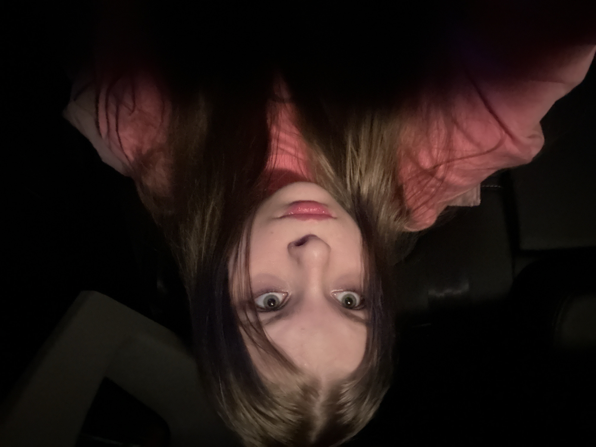 A young person is pictured upside down in what appears to be a dimly lit interior space. The person has long brown hair with some purple streaks and is wearing a pink shirt. The background is dark, with glimpses of what might be a car interior or some other enclosed space. The location is not identifiable as Moscow, Russia from the available visual information.