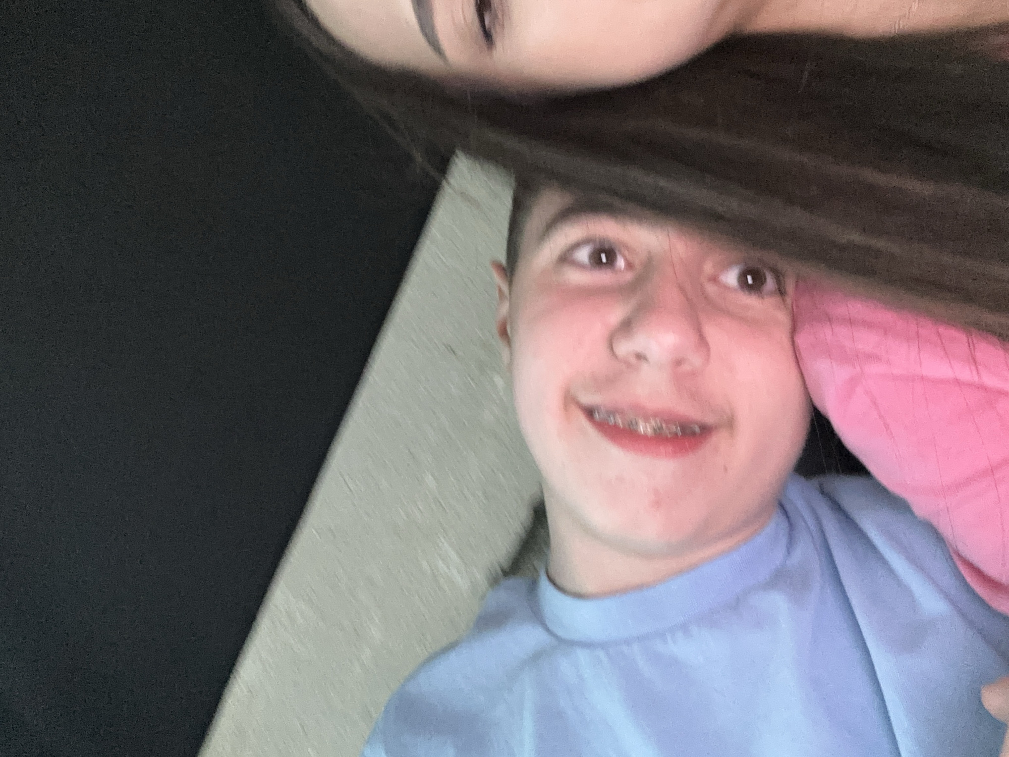 The image shows two people in close proximity. One person, with dark hair, is partially visible in the top of the frame. The second person, wearing a light blue shirt, is smiling and looking upwards. They have braces on their teeth. Part of a pink fabric is visible on the right side of the frame, near the second person's face. There is a gray surface adjacent to the person in the blue shirt. A dark area occupies the left side of the frame. The location is unknown.
