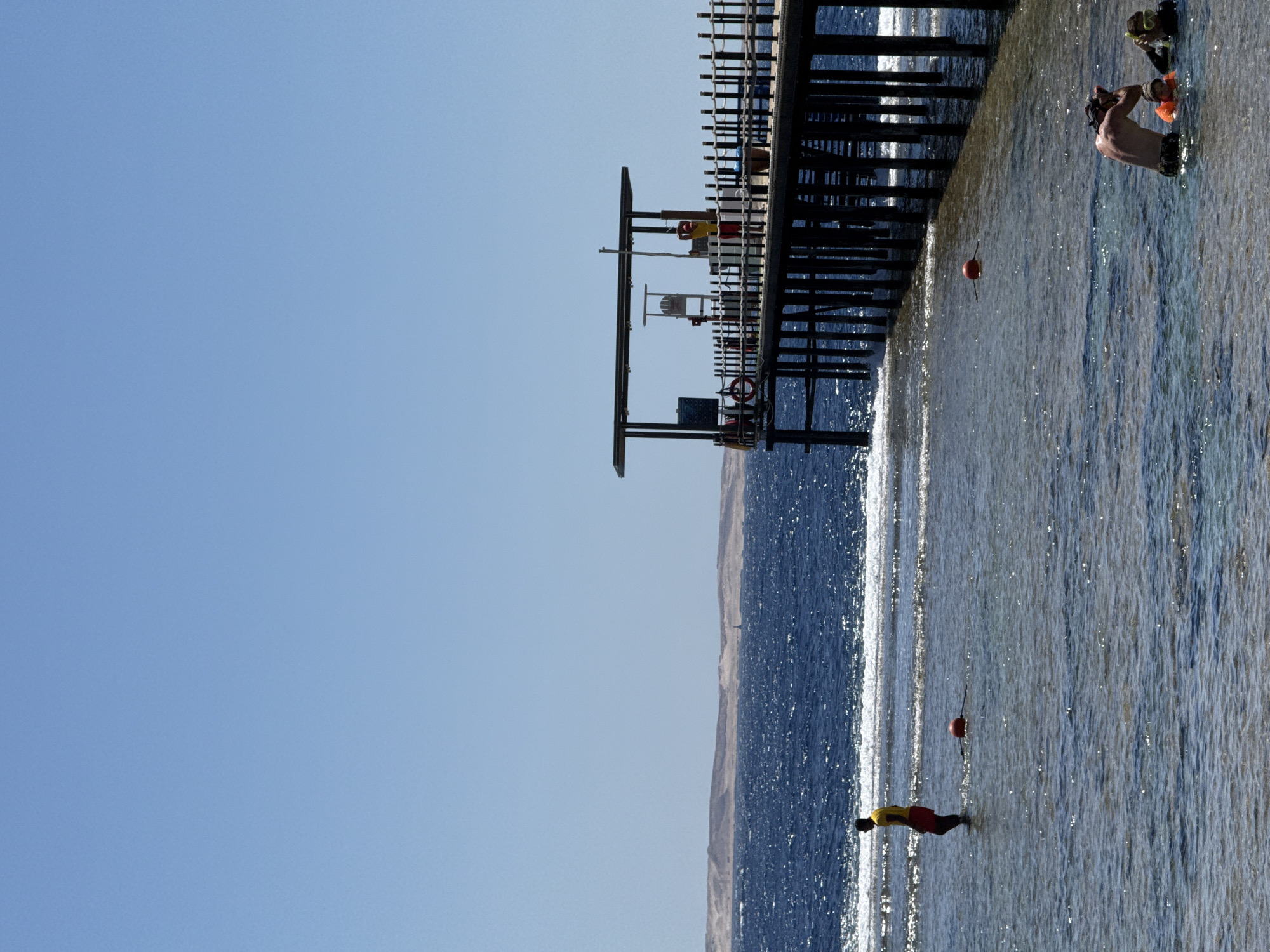 The image captures a tranquil scene at a pier, likely located in Sharm El Sheikh, Egypt, under a clear blue sky. The pier, constructed with dark wooden posts, extends into the deep blue sea, with a covered structure at its end. A lifeguard in a yellow top and red shorts stands watch in the shallow, clear water near the shore, observing the sea. Further out, a few individuals are engaged in water activities, one with a snorkel and mask, and another wearing a life vest. Red buoys are visible, marking the boundaries of the swimming area. The background reveals a distant coastline with undulating sandy hills. The overall atmosphere is one of leisure and safety, characteristic of a resort town like Sharm El Sheikh.