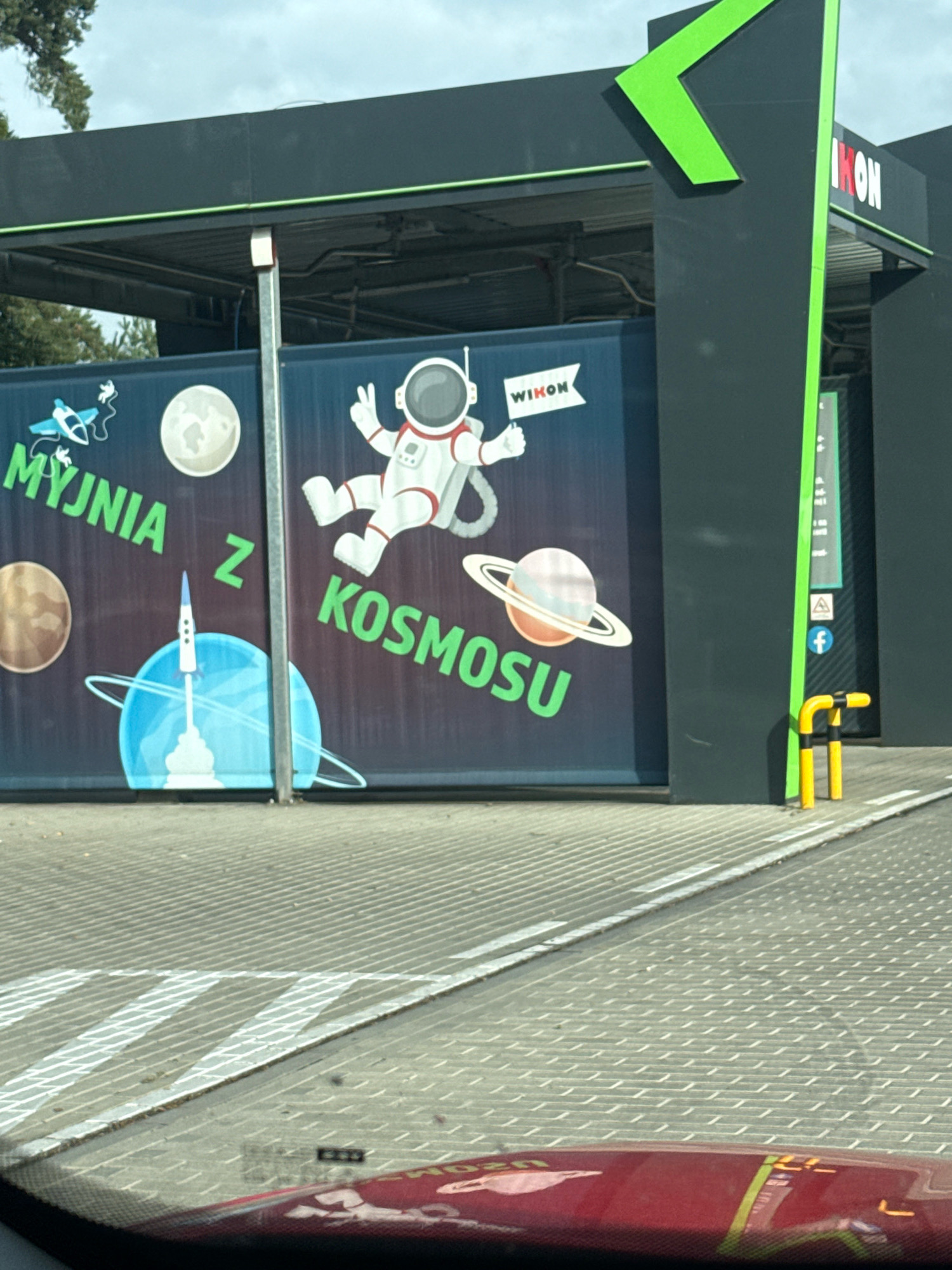 The image depicts a car wash facility with a prominent mural on its exterior wall. The mural features a space-themed design, including an astronaut, planets, and a rocket. The text on the mural, in green lettering, reads "MYJNIA Z KOSMOSU," which translates to "Car Wash from Space." There is also a white banner with the word "WIKON" printed on it, held by the astronaut. The car wash structure itself is dark grey with bright green accents. In the foreground, the reflection of a red car's dashboard is visible on the windshield. The ground in front of the car wash is a paved surface with a patterned texture and white markings. The location is identified as Rudno, Poland, on the exterior of a Wikon car wash.