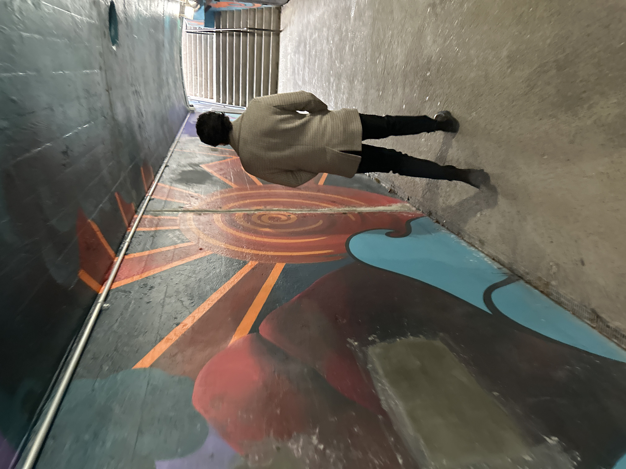 A person is depicted walking or standing in a vibrantly muraled pedestrian tunnel or underpass, located in the Village of Cold Spring, USA. The image is presented from a tilted perspective, giving the impression that the ground is a wall. The central figure is seen from behind, wearing a light, textured, perhaps checkered, long coat and dark trousers with dark shoes. They face away from the viewer, moving towards a brighter area at the end of the passageway.
The floor of the tunnel is covered in colorful street art, dominated by a large, stylized sun motif with radiating orange and red rays. This is complemented by abstract shapes in shades of blue, dark red, and black, which extend across the ground. The left wall of the tunnel is a dark, textured concrete, featuring a small, circular opening. A metal railing runs along the base of this wall, extending towards the background where a set of stairs or a ramp with further railings is visible, suggesting an exit or transition point. The right wall is a lighter, textured concrete surface. The lighting within the tunnel appears even, indicating an indoor or covered setting during daytime, or consistent artificial illumination. No specific activity beyond walking or standing is discernible, nor are any emotions conveyed. There is no visible text in the image.