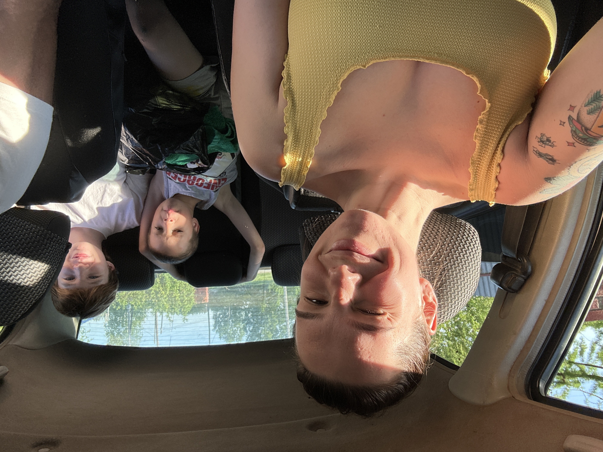A photograph taken inside a car shows three people from a low angle looking upward toward the camera. The leftmost person, a child with reddish-brown hair, is wearing a white shirt. A second child, also wearing a white shirt, is positioned next to the first. Above them is the adult taking the photo wearing a yellow tank top. A tattoo is visible on the adult's upper left arm. A black plastic bag is also visible. Through the car windows, trees and possibly a fence or net are visible. The location is Альняш, Russia.