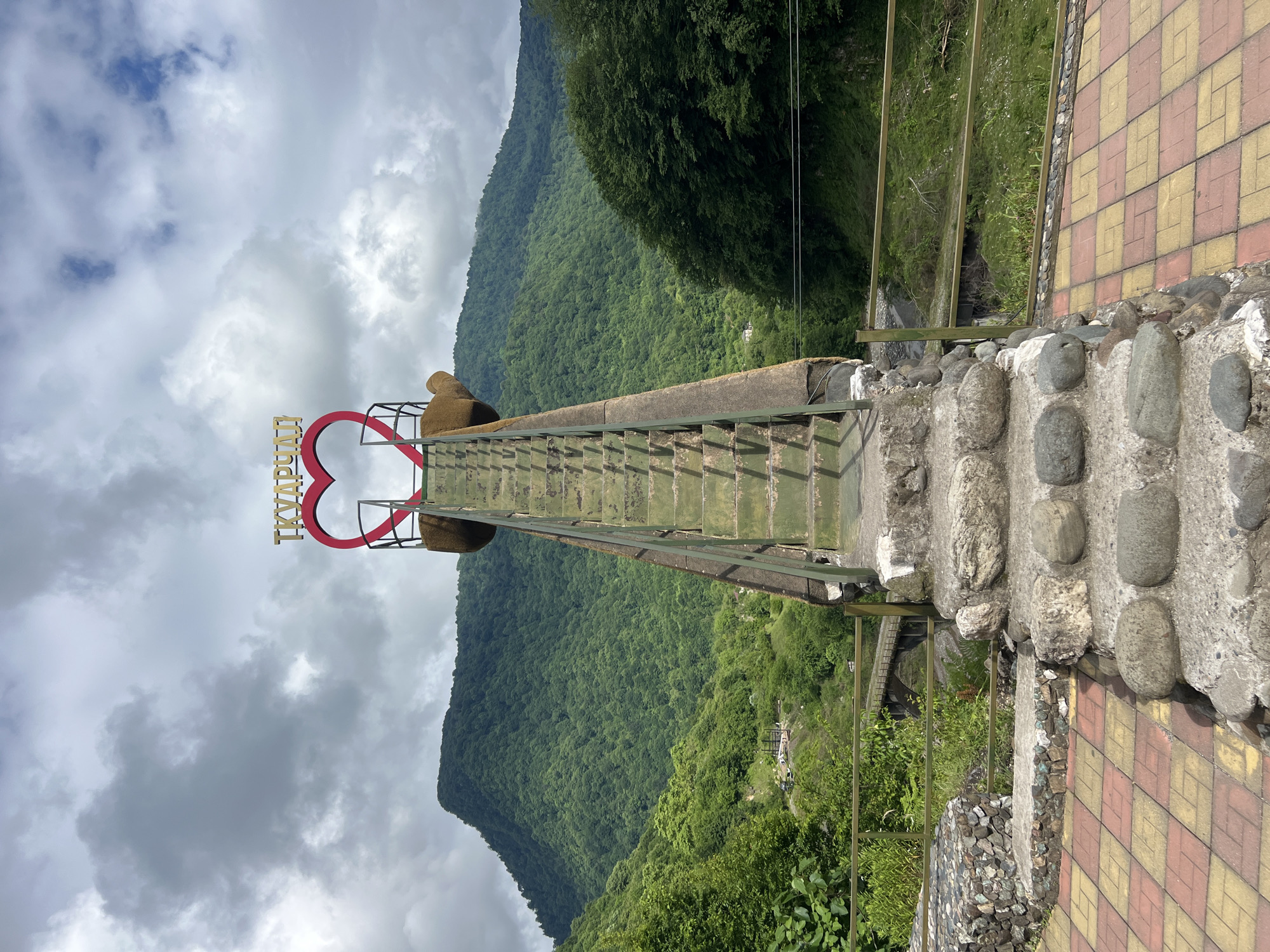 A wide, overcast shot shows a monumental, stone and metal stairway leading up to a large, red heart-shaped archway. The archway is adorned with gold lettering that reads "Ткварчалі". The stairway is weathered, with green metal railings and concrete steps. The heart arch is situated atop a pedestal that resembles a stylized bear paw.
The stairway is positioned in a scenic, mountainous region. Lush green trees and foliage cover the steep slopes of the mountains in the background. A river or stream runs through the valley below, spanned by a stone bridge in the mid-ground. The foreground features a paved area with red and yellow square tiles and a stone retaining wall.
The lighting suggests an overcast day, with diffused sunlight. There are no people visible in the image. The overall scene conveys a sense of a tourist attraction or viewpoint in a natural, rural setting. The lettering on the arch indicates that the location is likely Tkvarchal or Tkuarchal, a city in Abkhazia.