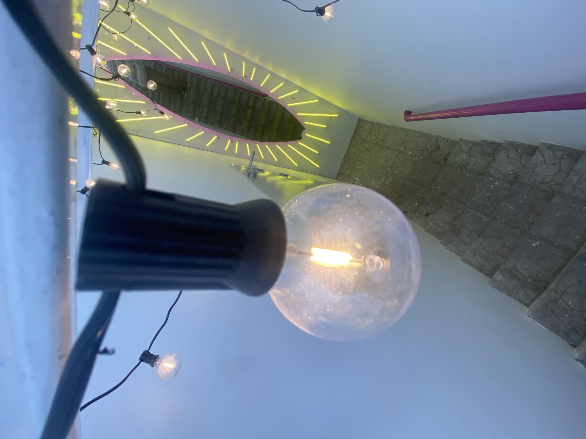 A downward-angled shot captures a string of illuminated light bulbs in the foreground, hanging from a dark cord that stretches across the top of the frame. The largest bulb, in sharp focus, glows warmly, its filament visible. Behind it, a narrow staircase with grey, textured steps descends into shadow. To the right, a wall is adorned with a surfboard-shaped mirror outlined in bright yellow neon lights and a thin pink neon border. A person, reflected in the mirror, is seen from the chest down, wearing striped clothing and dark trousers. A pink handrail runs down the side of the staircase, bordering a pale blue wall. The overall impression is an interior space, possibly a hallway or staircase, decorated with eclectic lighting and a prominent mirror. The location in Khabarovsk, Russia is not discernible from the visual information provided in the image.