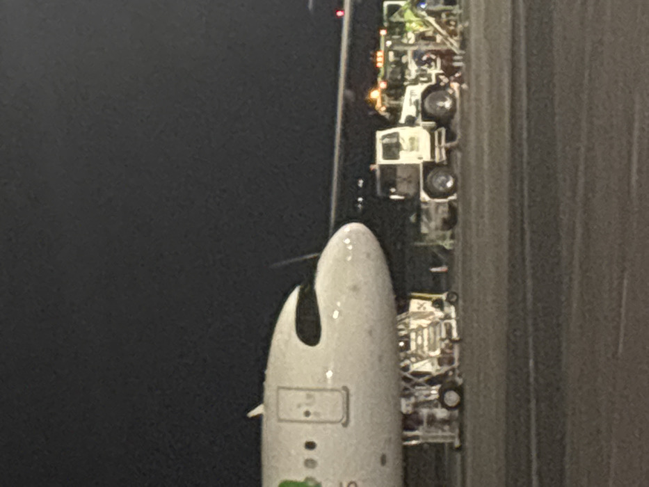 From an aerial perspective, the nose of a white aircraft is positioned adjacent to a dark tarmac. Next to the aircraft on the tarmac, various ground support vehicles are parked. The scene appears to be taken at night, illuminated by artificial lighting from the vehicles. The rest of the image is divided between the dark sky and the texture of the tarmac.