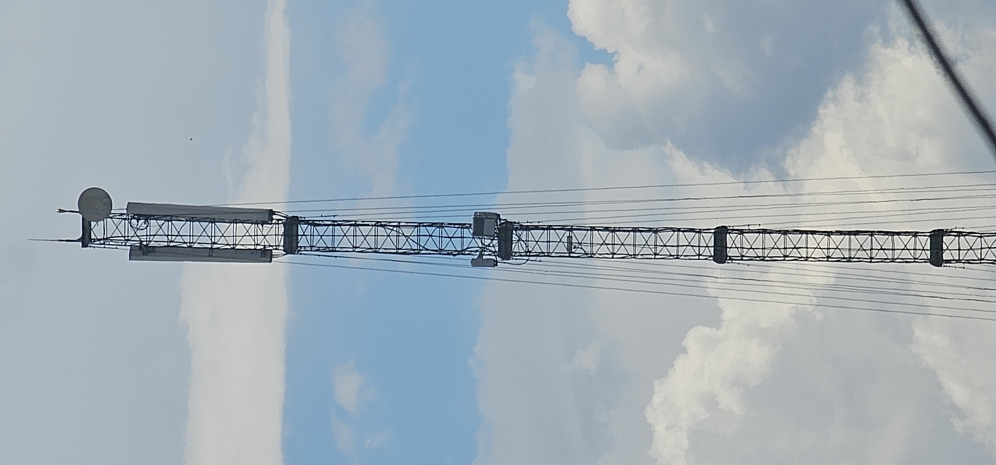 This is a vertical, medium shot of a tall, lattice telecommunications tower against a cloudy sky. The tower is constructed of dark metal beams forming a narrow, vertical grid. Attached to the tower are several antennas and equipment boxes. Prominently visible are two large, rectangular panels on opposite sides of the upper section of the tower, and a round dish antenna near the very top, along with a thin, vertical rod antenna. There are multiple cables running down the sides of the tower. The sky is a mix of pale blue and white clouds. There are no people or vehicles visible in the image. The scene appears to be an outdoor environment, characteristic of a telecommunications infrastructure site. The image does not provide any specific information that would allow for a precise location within Puncak Jaya, Indonesia, beyond it being a telecommunications tower in that region.