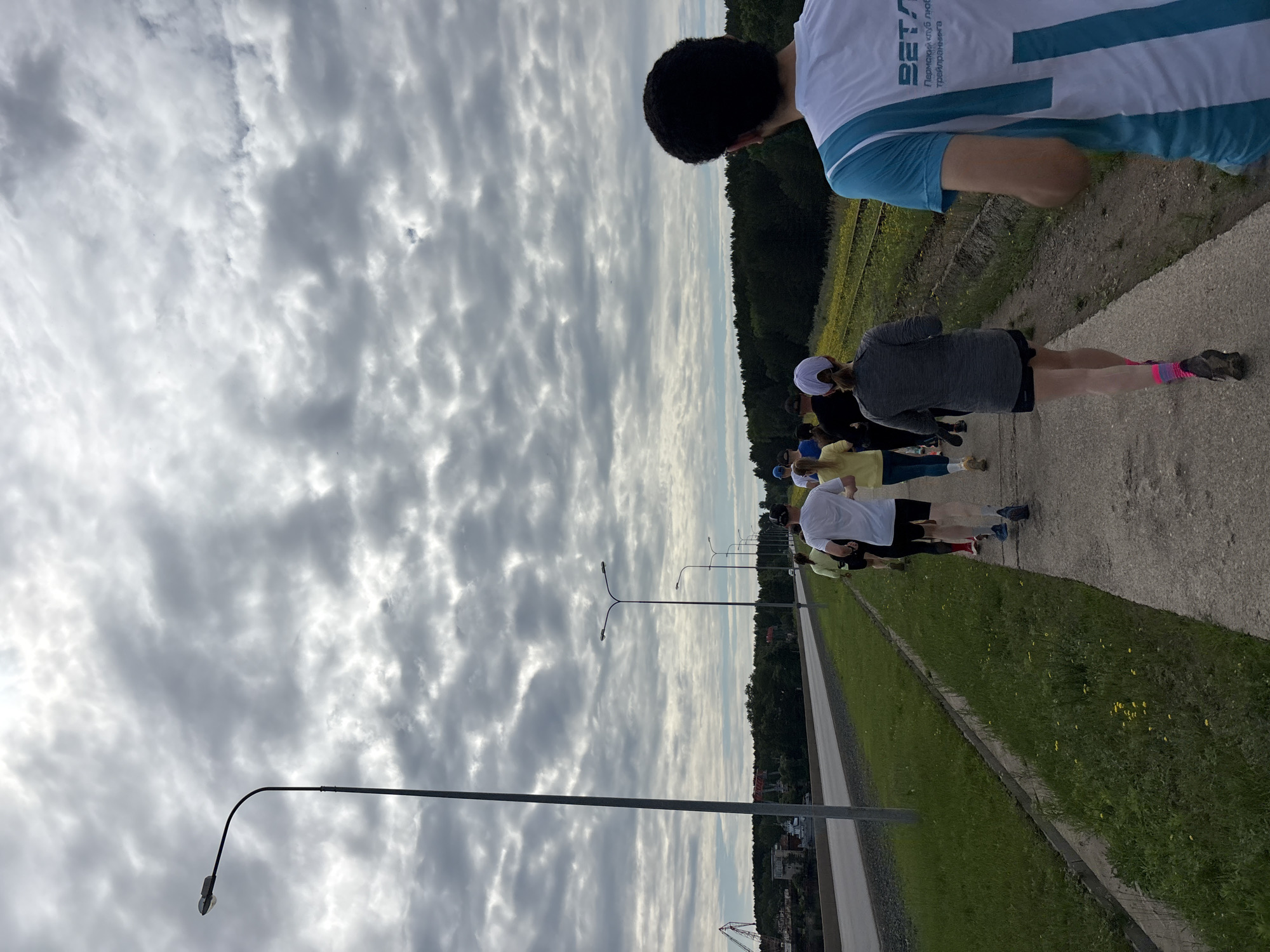 The image captures an outdoor scene in Chaikovsky, Russia, featuring a group of individuals running on a paved pathway under a heavily overcast sky. The sky is dominated by dense, textured grey clouds, with brighter areas indicating diffused daylight.
The scene is bisected horizontally by a paved multi-lane road running parallel to the pathway. To the left of the road, beyond a grey barrier, an industrial or port area is visible, characterized by large red-and-white and grey industrial cranes, various structures resembling buildings or ships, and a backdrop of dense green foliage, likely indicating a waterfront or riverbank. A tall, single-armed grey streetlight stands prominently on the far left, near the road.
To the right of the paved pathway where the runners are, there is a narrow grassy verge containing small yellow flowers, followed by two parallel railway tracks. Beyond the tracks, a dense line of dark green trees forms a forest or wooded area, extending into the background. Several double-armed streetlights are spaced along the right side of the road, illuminating it.
Approximately seven to ten individuals are depicted running away from the viewer along the concrete pathway. In the immediate foreground on the right, a male runner is visible from the back, wearing a white and light blue technical running shirt. The back of his shirt features the logo "БЕТА" and the Russian text "Пермский клуб любителей трейлраннинга," translating to "Permian Club of Trail Running Enthusiasts." He has dark, curly hair. Ahead of him, a female runner wears a grey long-sleeved top, black shorts, a white baseball cap, and distinct bright pink socks. Other runners in the group are dressed in typical athletic wear, including white and yellow t-shirts, shorts, and leggings, and are engaged in the act of jogging or running along the route.