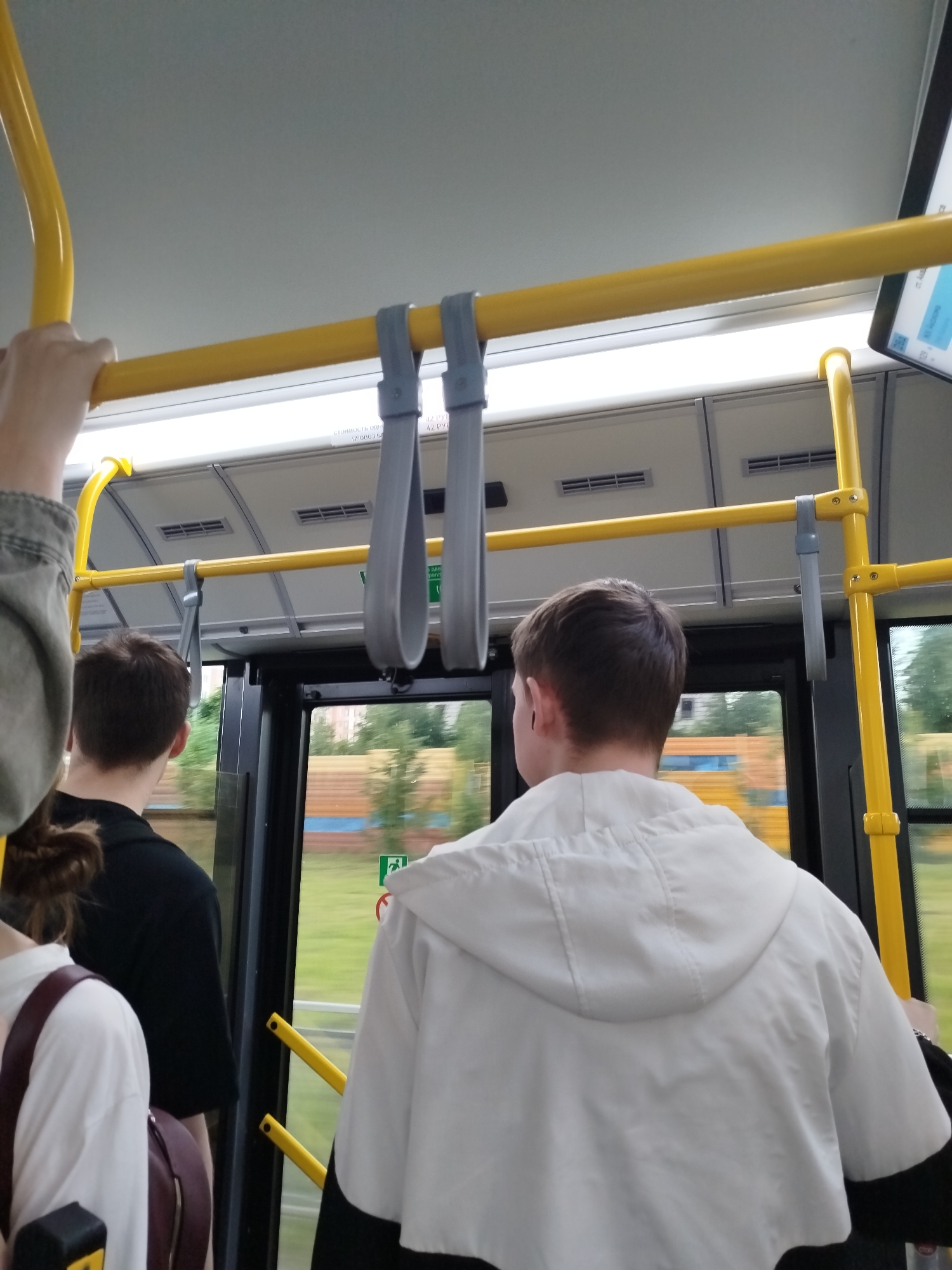 The image captures the interior of a bus in Yekaterinburg, Russia, focusing on passengers and the bus's internal structure. In the foreground, the back of a young man wearing a white hooded jacket is visible. To his left, another passenger in a black t-shirt is seen from the back. A person's arm, wearing a grey denim jacket, is visible on the far left. The bus features yellow handrails and grey hanging straps for passengers. Through the large windows, a blurred view of trees and buildings suggests the bus is in motion. The overall scene depicts a typical commute on public transportation in Yekaterinburg.