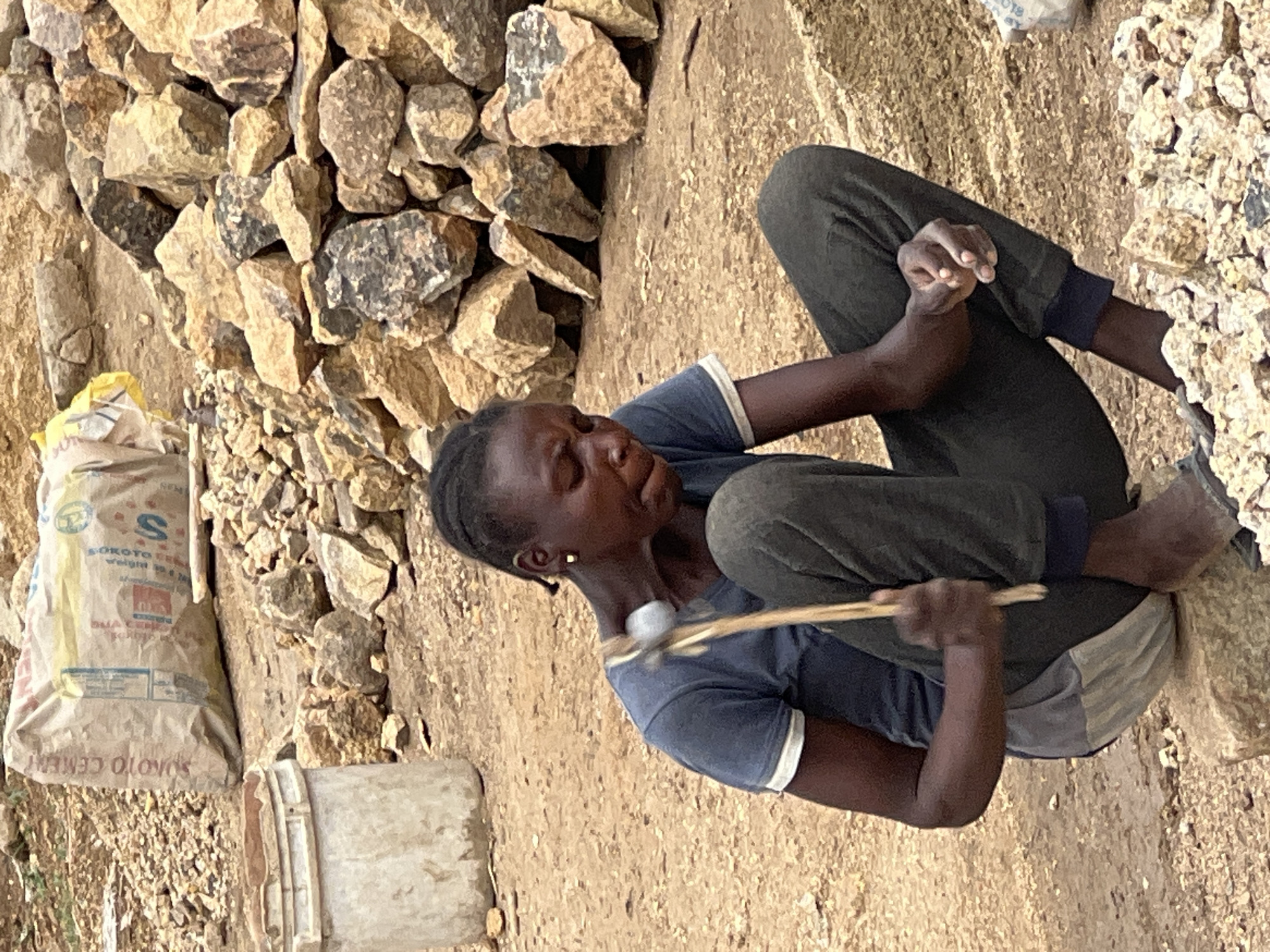 The image captures a young person, likely a child or young adult with dark skin and braided hair, engaged in manual labor outdoors. The individual is sitting on the dusty ground, barefoot, wearing a dark blue short-sleeved t-shirt with white trim and dark trousers. They are hunched over, focused intently on breaking rocks. In their hands, they hold a simple tool – a small, light-colored hammer-like object attached to a wooden stick – which they are using to strike a rock.
The setting appears to be a quarry or a site for processing raw materials, with numerous rocks and broken stones scattered across the light brown, sandy ground, forming piles around the worker. To the left of the person, a white sack stands upright, identified by the visible text "SOKOTO CEMENT" and "50 kg," with a partial "DANGOTE" logo also discernible. A light-colored bucket is placed near the sack.
The scene is illuminated by bright, direct sunlight, indicating daytime and clear weather. The person's expression is one of concentration and effort, with their eyes cast downwards on their task. The activity suggests arduous work, possibly involving the preparation of aggregate for construction. The location context is Gura Top, Nigeria.