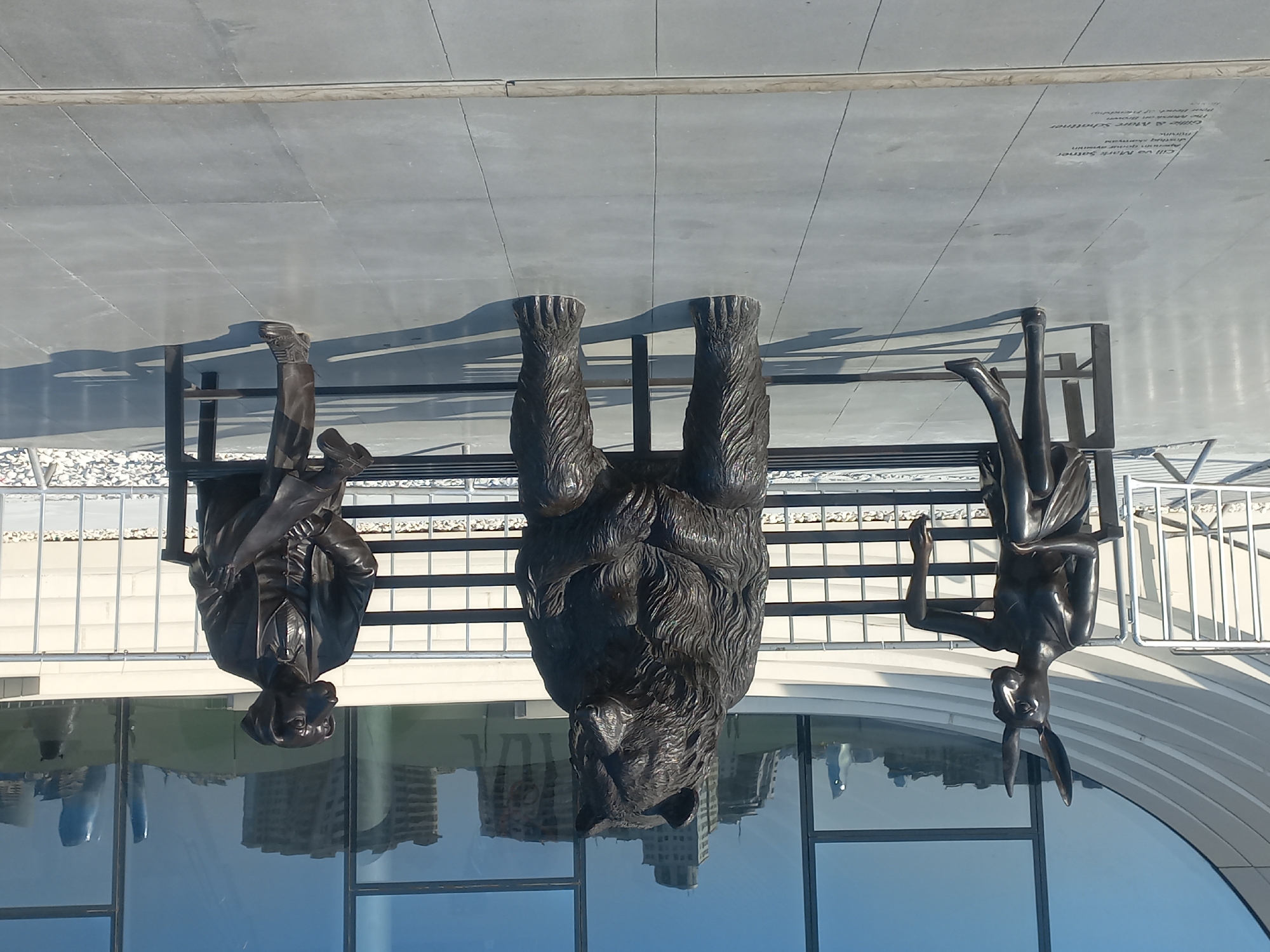 The image shows a public artwork in Baku, Azerbaijan, featuring three bronze statues of anthropomorphic animals sitting on a bench. From left to right, there is a rabbit woman, a bear, and a dog man dressed in a suit. The statues are positioned in front of a modern building with large glass windows, which reflect the surrounding cityscape. The ground is paved with large gray tiles. In the foreground, there is an inscription on the pavement, indicating the artwork's title and artists, "Cill ve Mark Sattner" and "The Baron on Brown Bench of Friendship". The setting appears to be a plaza or promenade, with a white metal railing in the background. The sky is clear and blue, suggesting a sunny day.