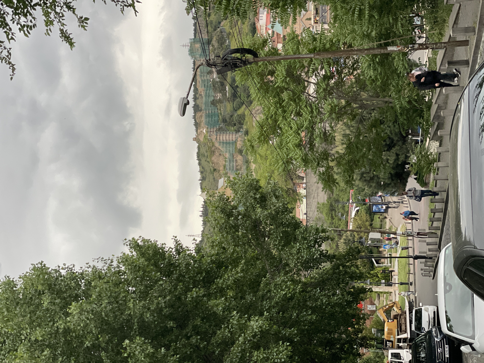 The image presents a street scene in Tbilisi, Georgia, captured on an overcast day. The perspective is slightly elevated, likely from a higher floor or hill, looking down onto a street that slopes gently downhill.
In the foreground, a mix of parked cars, primarily black and white, line the right side of the street. A small yellow CAT excavator is also parked alongside.
The street contains pedestrians walking. A crosswalk is visible. A couple walks together on the sidewalk on the right-hand side, hugging each other.
Beyond the street, the landscape ascends into a hillside covered with trees and buildings. A partially constructed or renovated building stands out due to extensive green scaffolding. Electrical poles and wires are present, adding to the urban landscape.
The sky is overcast, covering the city in a muted light. Overall, the image depicts a typical urban scene with a mix of infrastructure, vehicles, and pedestrians in Tbilisi.