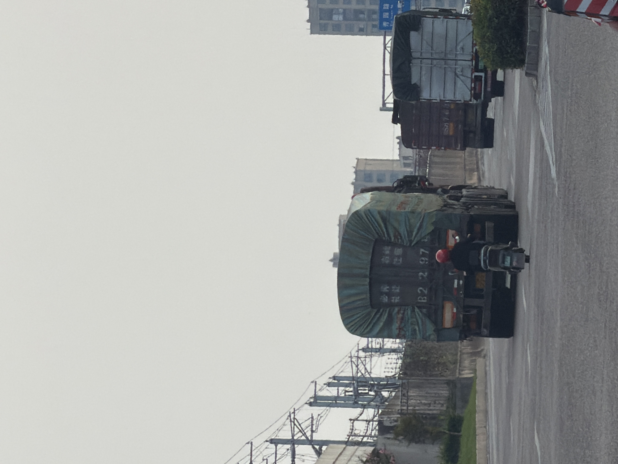 In the city of Jiangjin, China, on a road with multiple lanes, a motorcyclist wearing a red helmet is riding a small electric scooter behind a large green-covered truck. The truck has Chinese characters and the license plate "B2 297" visible on its rear. To the right of this truck, another truck with a silver-colored cover is also visible, and further to the right, a dark-colored truck is seen. In the background, tall buildings and electric power lines with poles are present. The sky is overcast.