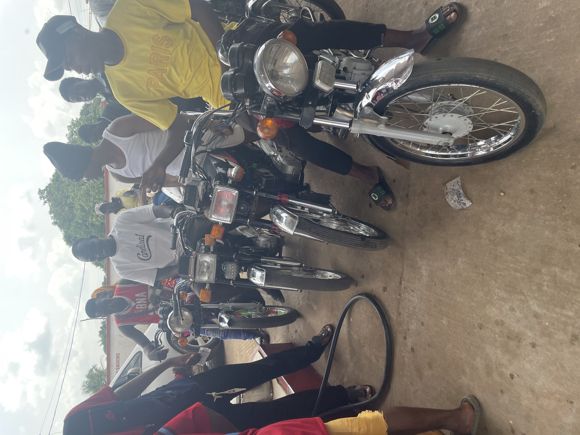 The image shows a group of people gathered around several motorcycles. The people appear to be predominantly Black African men, dressed in casual clothing. Some are sitting on the motorcycles. The motorcycles are various models, likely common in Nigeria. The setting appears to be a paved or concrete area, possibly a roadside or a small business. A motorcycle tire lies on the ground. The scene is located in Akwanga, Nigeria.