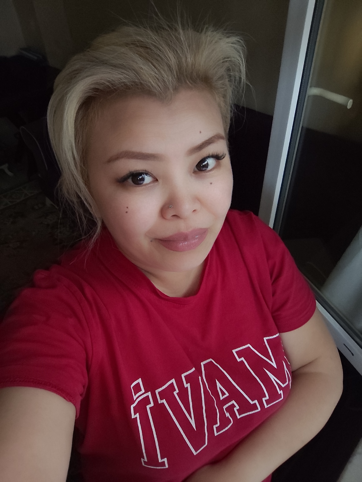 A head and shoulders shot captures a woman with light blonde hair. She is wearing a red t-shirt with the name "IVAN" printed on it in large, white letters. The woman has fair skin, dark eyes, and a small nose ring. There are two small moles on her left cheek. Her makeup includes eyeliner and lipstick. The background is slightly blurred but appears to show a window frame on the right and dark, indistinct shapes of furniture on the left. The location is Unknown, Unknown.