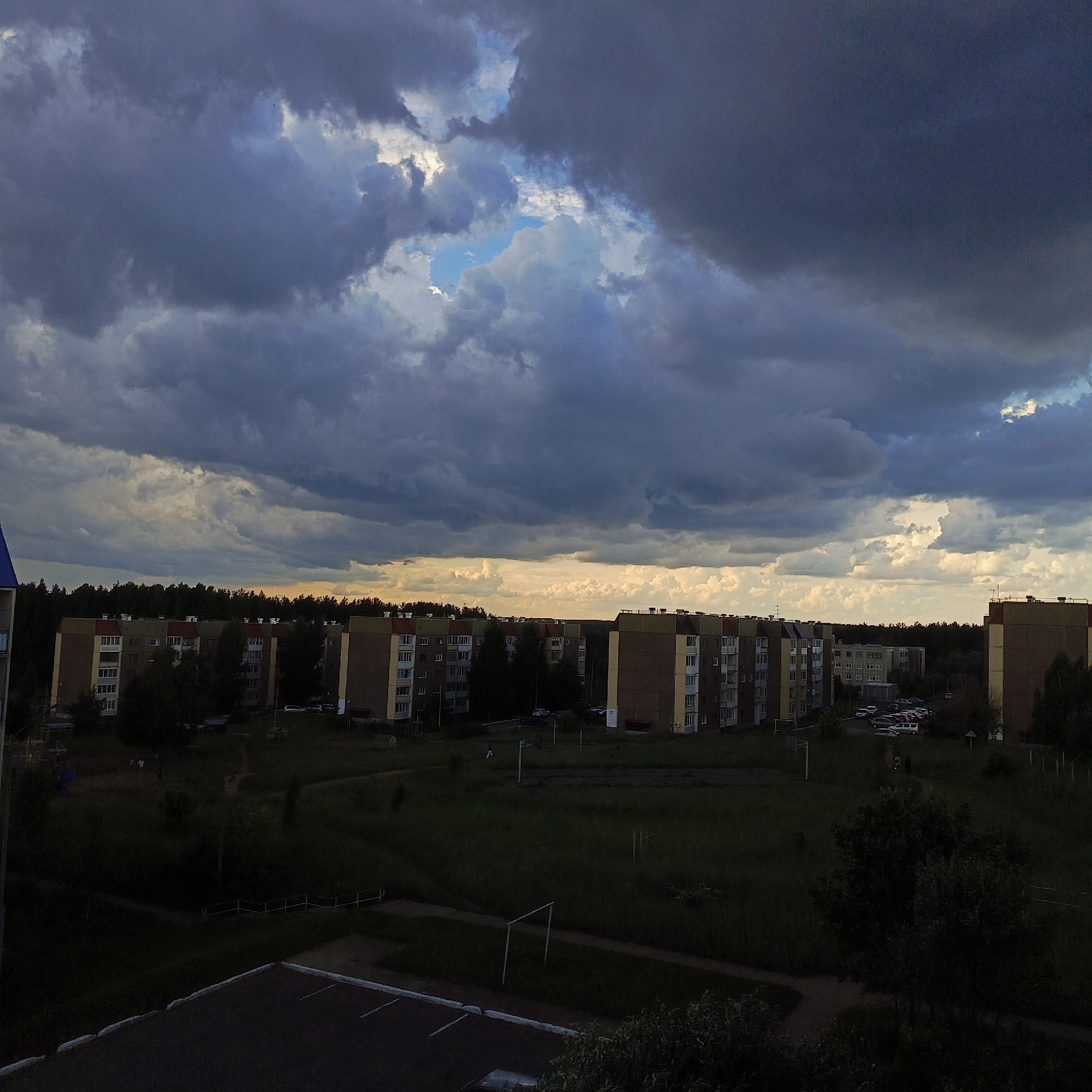 The image shows an exterior view of a residential area in the city of Markovsky, Russia, under a dramatic, cloud-filled sky. In the foreground, a dark, paved surface with white markings, likely a parking lot or road, occupies the bottom portion of the frame. Beyond this, a grassy area with scattered trees and a children's playground can be seen. Several multi-story apartment buildings, characterized by their beige and red facades, are visible in the midground, stretching horizontally across the scene. A dense line of trees borders the horizon behind the buildings, and a sliver of bright, yellow light suggests the setting sun peeking through the clouds. The sky above is dominated by dark, heavy storm clouds, with patches of lighter grey and a small opening revealing a bright blue sky. There are a few indistinct figures walking in the grassy area. The overall impression is one of an impending storm in a suburban or semi-urban environment.