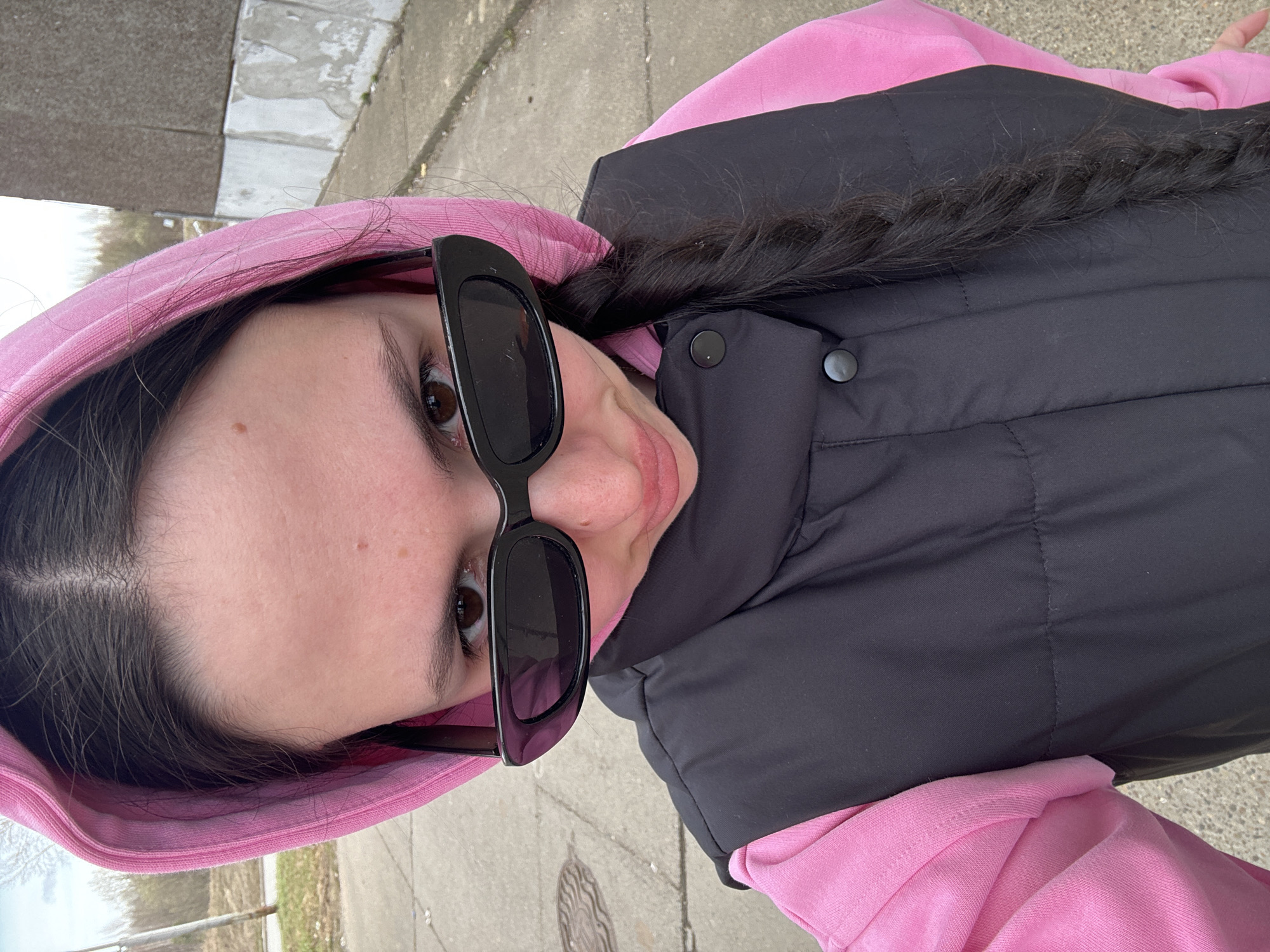 An overhead selfie captures a young person, likely female, outdoors in what appears to be Markovskiy, Russia. The image is rotated 90 degrees clockwise, showing the subject's head tilted to the right and the ground vertically. The person is looking directly up at the camera with a slight, subtle smile.
She is dressed in a bright pink hooded sweatshirt, with the hood up, covered by a black puffer vest. Her long, dark hair is styled in a prominent braid that rests over her left shoulder and against the black vest. She wears black, rectangular sunglasses pushed slightly down on her nose, allowing her brown eyes to be visible above the frames. A small mole is noticeable on her right cheek.
The background suggests an outdoor setting, likely a paved area such as a sidewalk or pathway, composed of light-colored concrete slabs. Further in the background, out of focus, are hints of green foliage or distant trees, indicating an open-air environment. The lighting suggests it is daytime, with a soft, even illumination that doesn't cast harsh shadows, possibly indicating an overcast sky. No visible text is present in the image.
