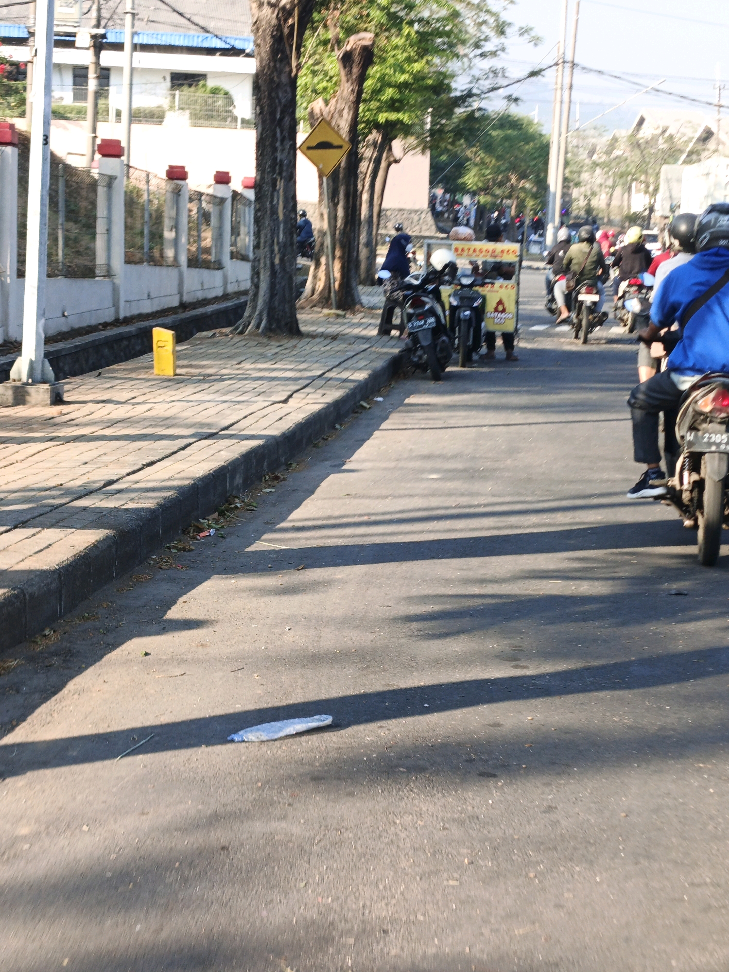 The image depicts a street scene in Ngoro, Indonesia, with a line of motorbikes on the right side of the road, some carrying riders. In the foreground, a clear plastic water bottle lies on the asphalt, partially illuminated by sunlight casting shadows from overhead trees. To the left, a sidewalk is visible, bordered by a gray concrete curb and lined with trees. A yellow post stands on the sidewalk. Further down the street, more motorbikes and pedestrians are seen, and a food cart with the word "BATAGOR" is prominent. Buildings and a fence are present in the background. The overall scene suggests a typical urban environment with active street life and traffic.