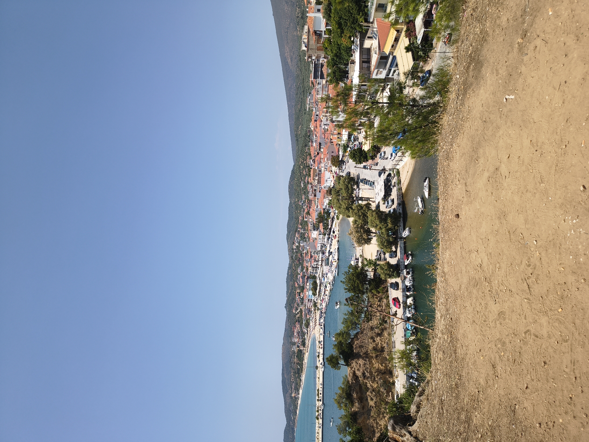 The image shows a panoramic view of Limenaria, Greece, on a clear, sunny day. The foreground is dominated by a sandy, barren hillside with scattered dry grass and pebbles. A path or track with visible footprints is present on the sandy surface.
Descending from the hillside, the midground reveals a small, sheltered harbor filled with numerous small boats and a few larger vessels docked. The water is a clear, greenish-blue. Along the edge of the harbor, a paved road runs parallel to the water, with several cars parked along it. Trees and vegetation line the harbor and the road.
Further into the background, the town of Limenaria sprawls across a gentle slope leading down to a sandy beach and the sea. Buildings with red-tiled roofs are characteristic of the architecture. A breakwater extends into the sea, with more boats visible in the bay beyond. The coastline stretches out to the left, with a sandy beach dotted with umbrellas and people. The background is formed by undulating, tree-covered hills. The overall impression is of a Mediterranean coastal town in a picturesque setting.