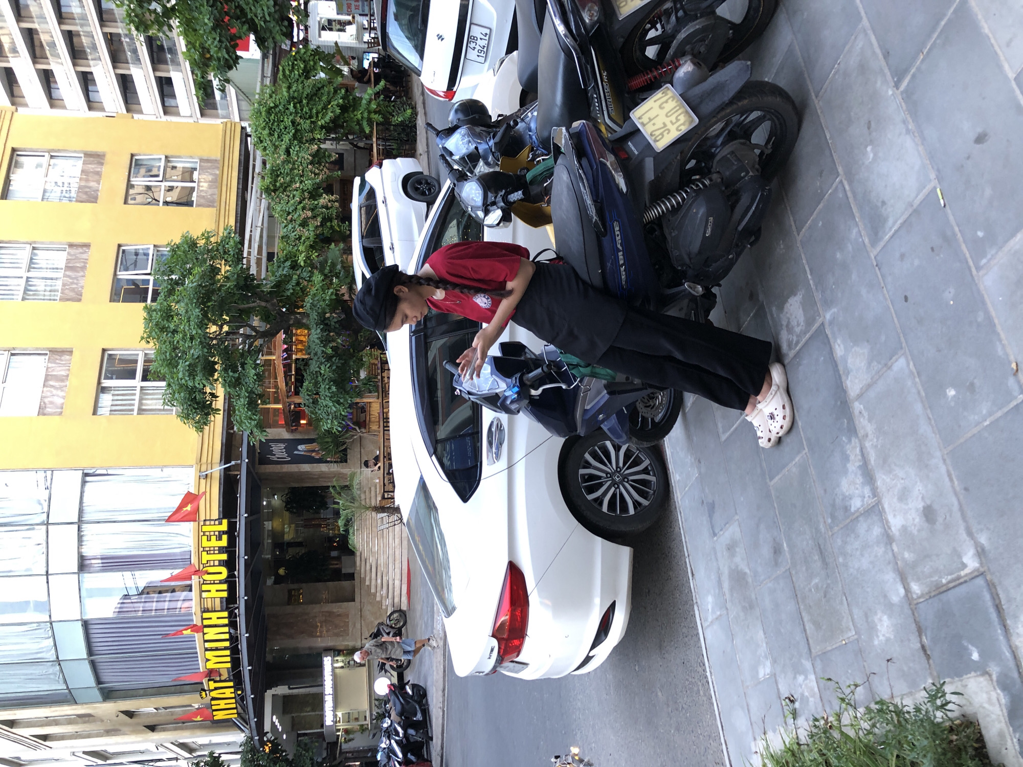 In this medium shot, captured in a street scene in Da Nang, Vietnam, a young woman with a braid is the central subject. She's wearing a red shirt and a black cap, with black pants and white clogs. Her attention is focused on her phone, which she holds in her hands while leaning against a parked white car. The car, with its taillights visible, is positioned alongside several parked motorcycles.
Behind the woman and the car, a building with yellow paint and multiple windows dominates the background, suggesting an urban environment. Red Vietnamese flags are displayed on the building. The ground is paved with gray tiles, and green foliage adds a natural element to the scene. The image appears to be taken during the day, with clear lighting. The overall atmosphere is casual and everyday, with no strong indications of a specific event, but the presence of parked vehicles and a building with a prominent sign hints at a commercial area. The sign on the building reads "NHAT MINH HOTEL".