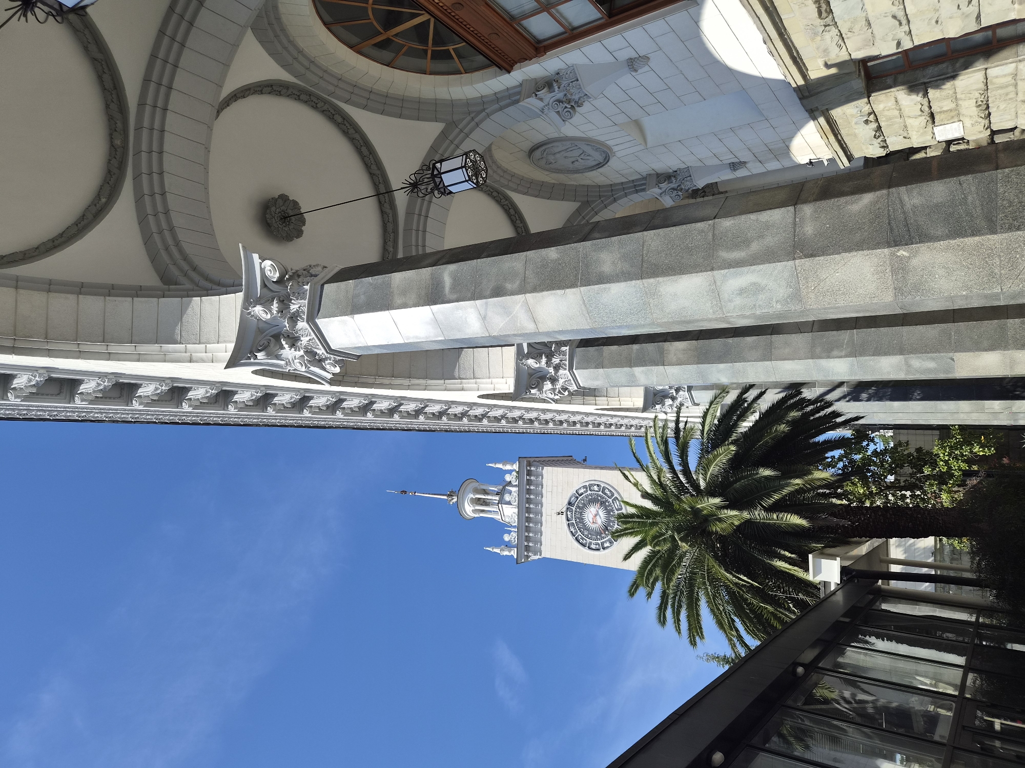 The image showcases a low-angle view upwards in Sochi, Russia, focusing on the architecture and a palm tree against a clear blue sky. The top portion of the frame shows the underside of a building's canopy with arched designs, decorative stone detailing, and an ornate lamp fixture. Below the canopy is a tower with a large clock face visible. A palm tree stands to the right of the tower. A modern glass structure is also partially visible in the lower right corner of the frame.