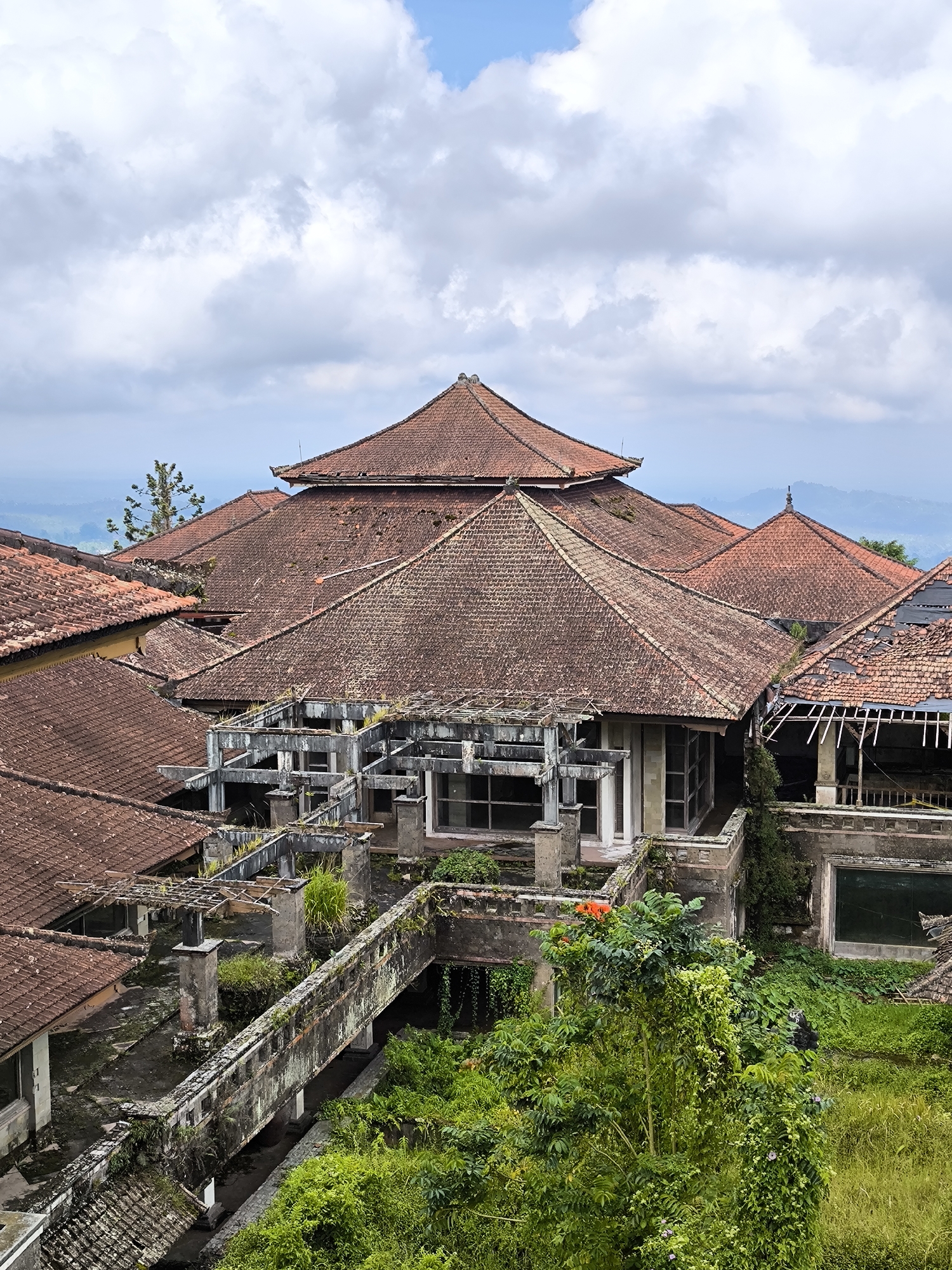The image depicts an aerial view of Taman Festival, an abandoned theme park located in Mekarsari, Indonesia. The structures are dilapidated and partially overgrown with lush green vegetation. Dominating the scene are the decaying roofs of various buildings, covered with reddish-brown tiles. A concrete bridge-like structure, heavily weathered and covered with moss and plant growth, extends into the lower section of the image. The skeletal remains of a building with a concrete framework and window openings are visible. A backdrop of distant hills is visible against a cloudy sky.