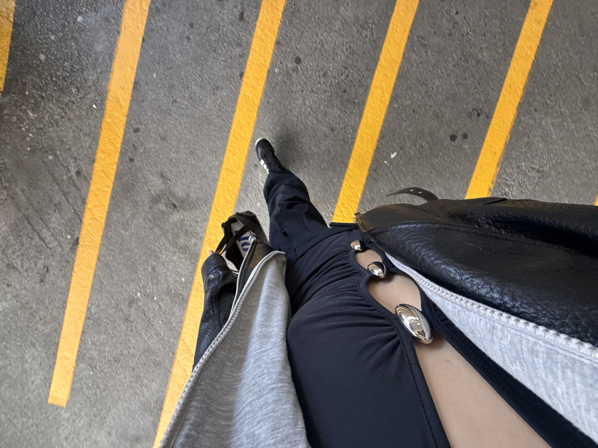 The photograph is a high-angle shot of a person standing on an asphalt surface with yellow parallel lines. The person is wearing black shoes, dark trousers, and a black dress with silver decorations. The person is also wearing a jacket. The setting is likely urban, based on the asphalt surface and yellow lines. The location is identified as Richmond, Canada, but without additional cues there is not enough information to provide any further specificity about a neighborhood, building, etc. in Richmond, Canada.