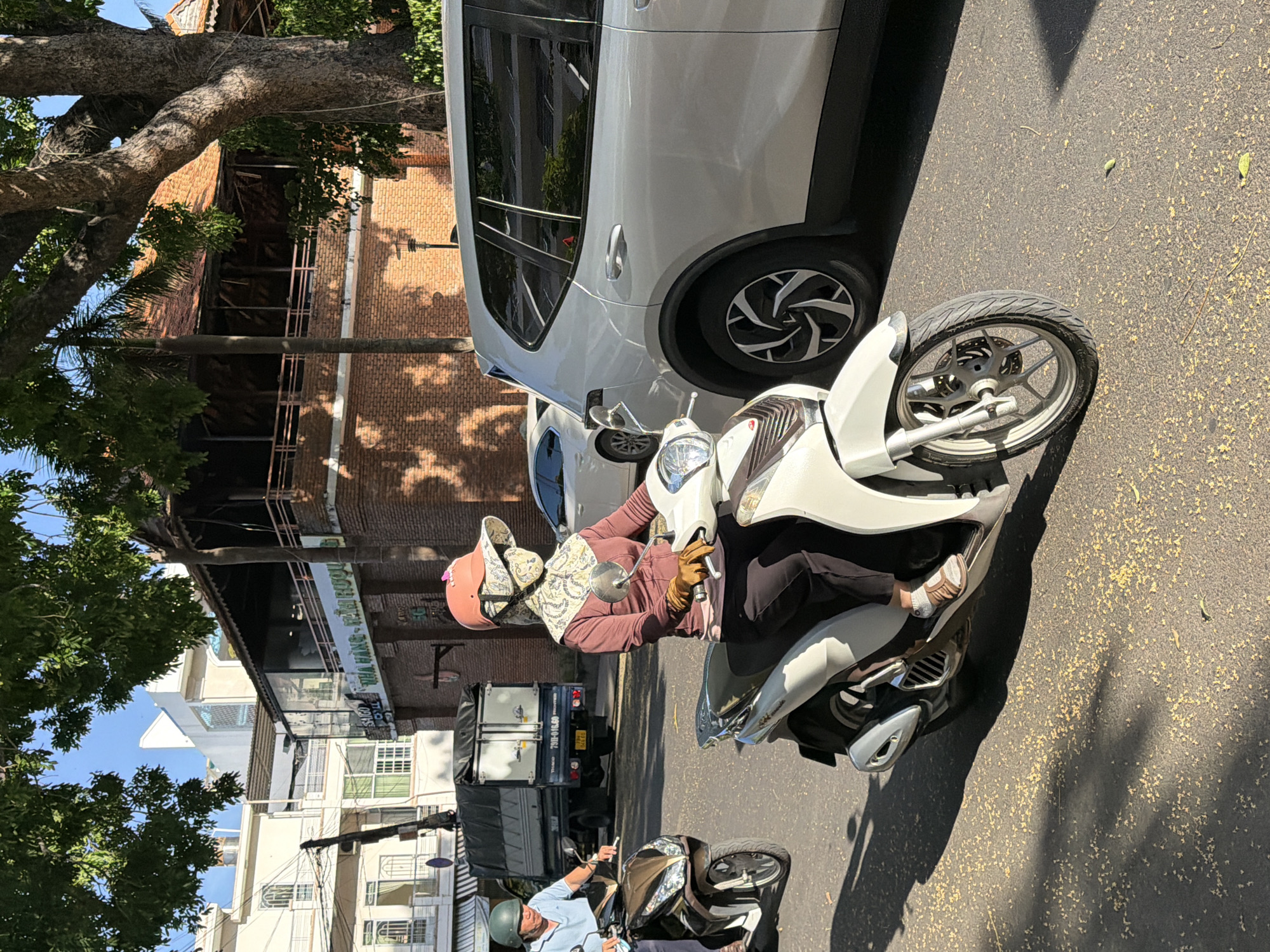 A woman wearing a face mask, hat, gloves, and a maroon jacket rides a white scooter in the foreground on a sunlit asphalt road in Nha Trang, Vietnam. The scooter is a Honda SH. She is on the right side of the road, facing away from the viewer. To her left, a man wearing a blue shirt and a green helmet rides a brown scooter. Behind them, a white truck with a grey tarp is parked. Further in the background, a white modern building and a traditional brick building with a wooden balcony are visible. The ground is covered with small yellow fallen leaves and shadows. The scene appears to be a street with vehicular traffic.