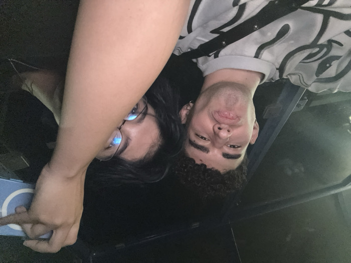 Two people are captured in an upside-down selfie. One person, likely female, wears glasses and has dark hair. The other, presumably male, has curly hair. Both are close to the camera. A gray and white patterned shirt with a black strap is visible on the right. Dark background surrounds them, with a glimpse of what appears to be a cell phone. They seem to be inside a vehicle, possibly a car.